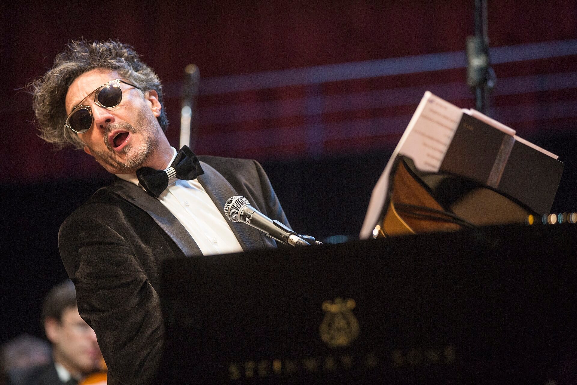 Fito Páez sings at a grand piano in a black tuxedo and dark round sunglasses, with sheet music open in front of him.