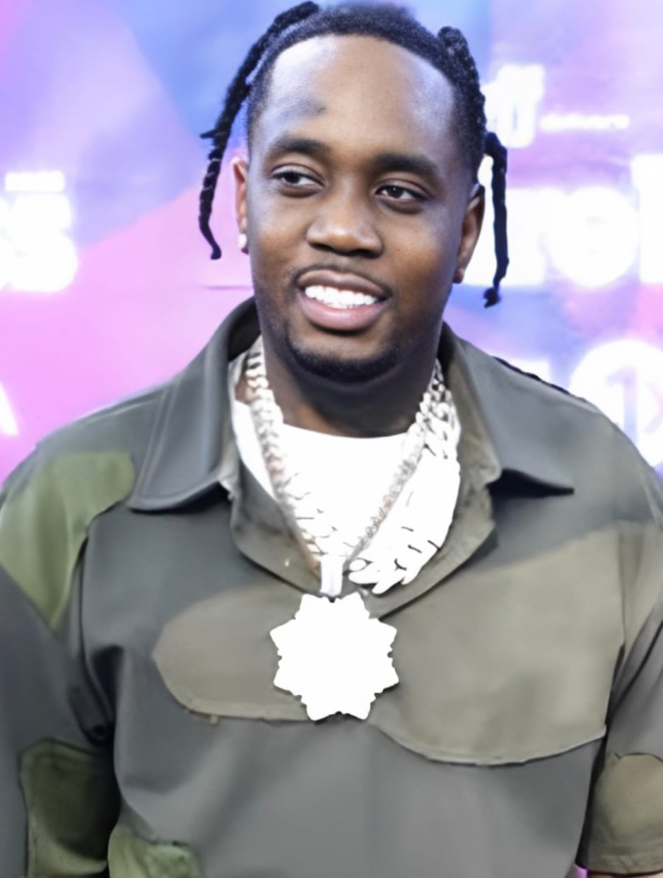 Fivio Foreign smiles in an olive green jacket and layered chain necklaces, with braided hair, in front of a purple event backdrop.