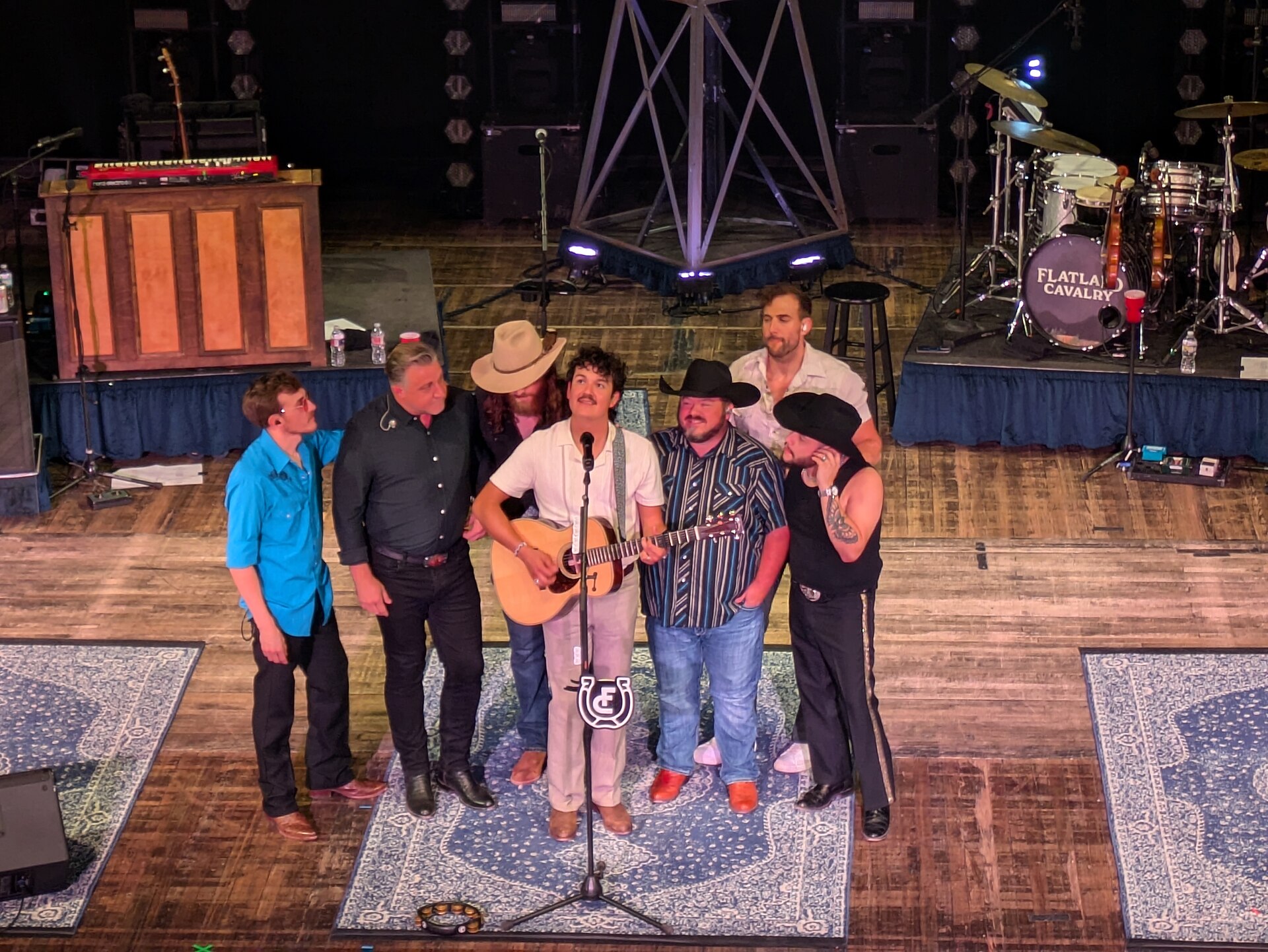 Flatland Cavalry play a theatre stage in red and amber light, the singer at a centre microphone with a fiddle player and guitarists spread across the stage.