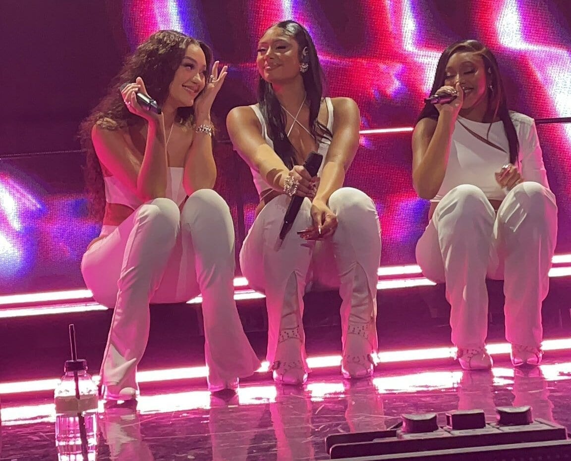The three members of FLO crouch on stage in matching white outfits under pink stage lighting, smiling and holding microphones.