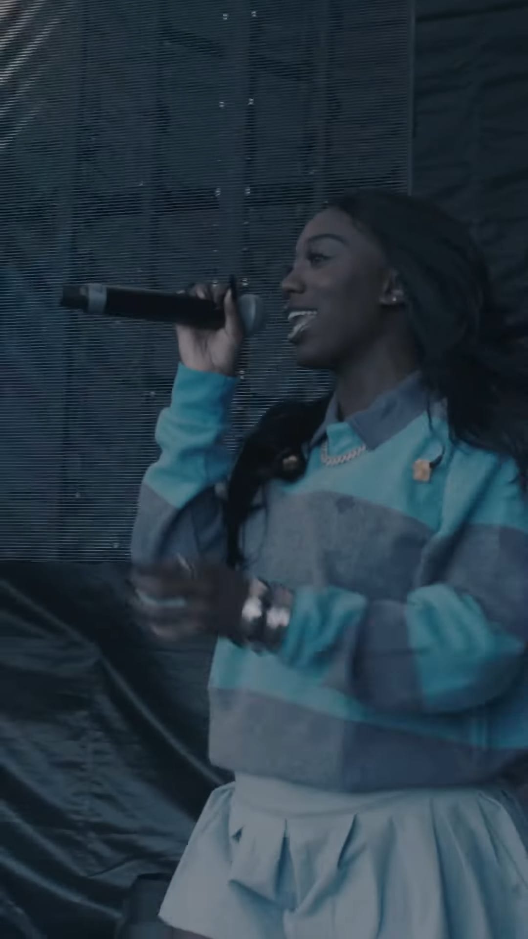 Flo Milli sings into a microphone against a shimmering blue mesh backdrop, wearing a teal and gray color-blocked top.