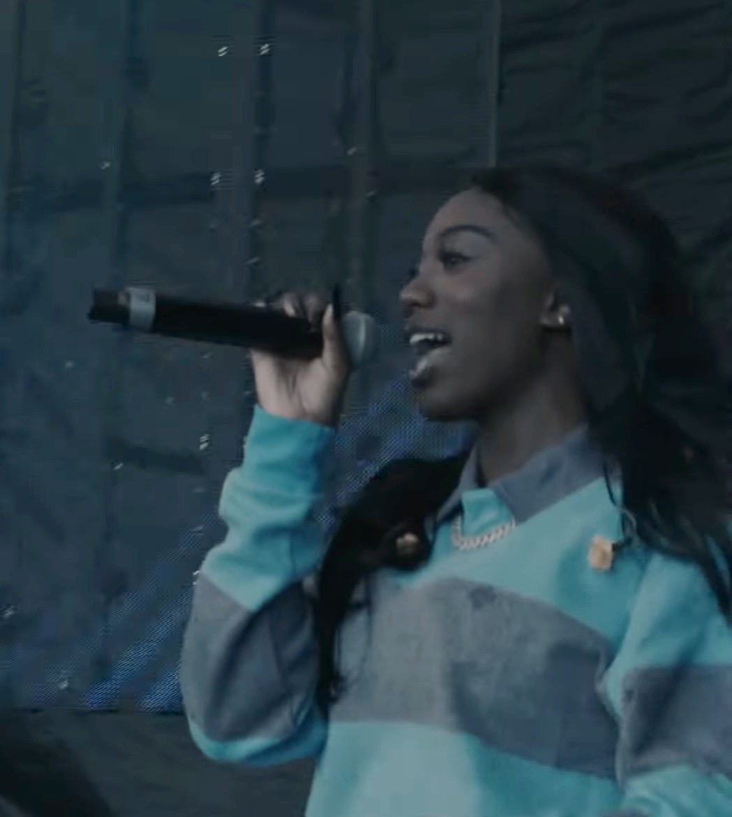 Flo Milli raps into a handheld microphone in profile, long braided hair down, wearing a blue and grey striped rugby shirt and a gold chain.