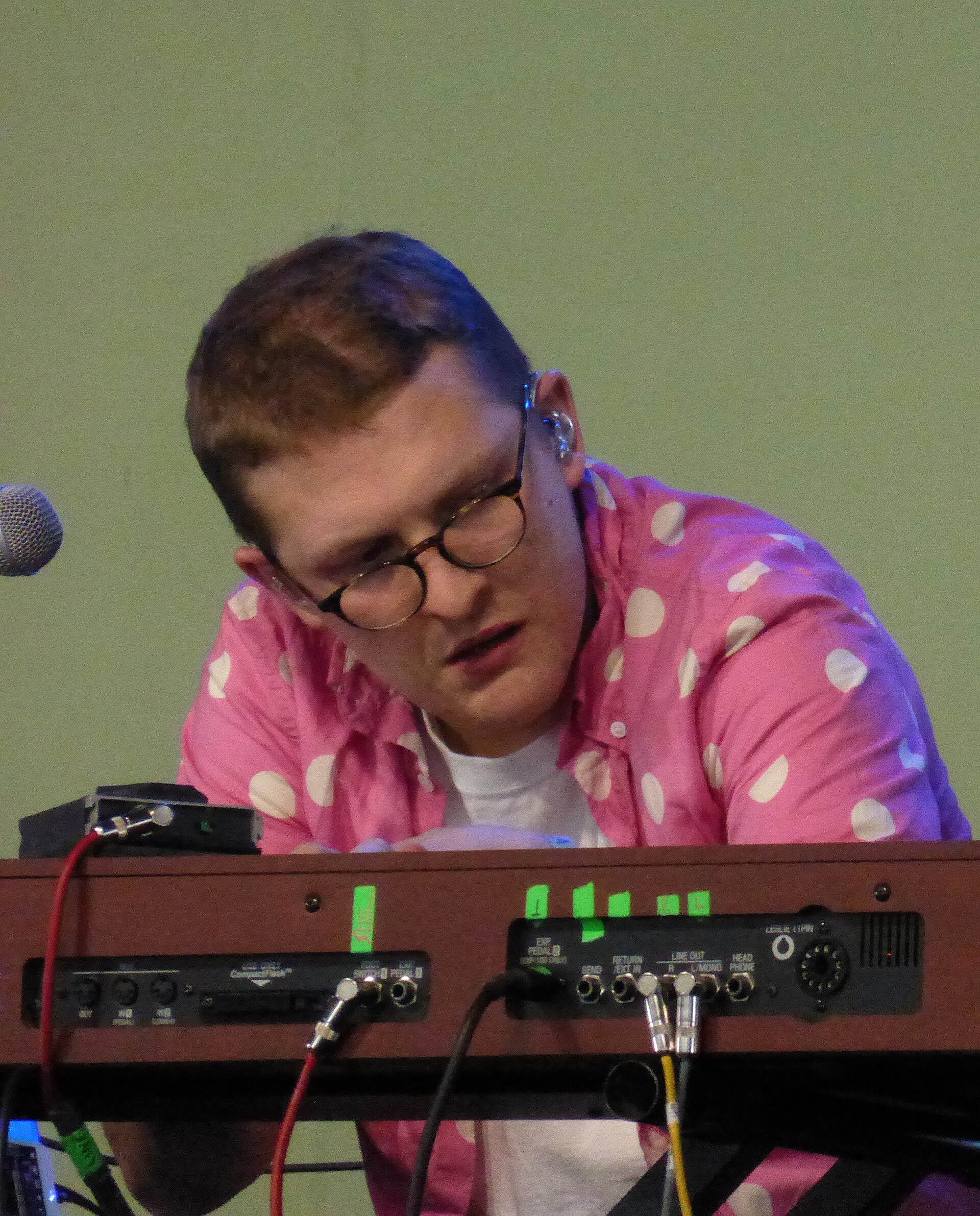 Sam Shepherd leans over a keyboard and a rack of outboard gear in a pink polka-dot shirt and glasses, mid-set.
