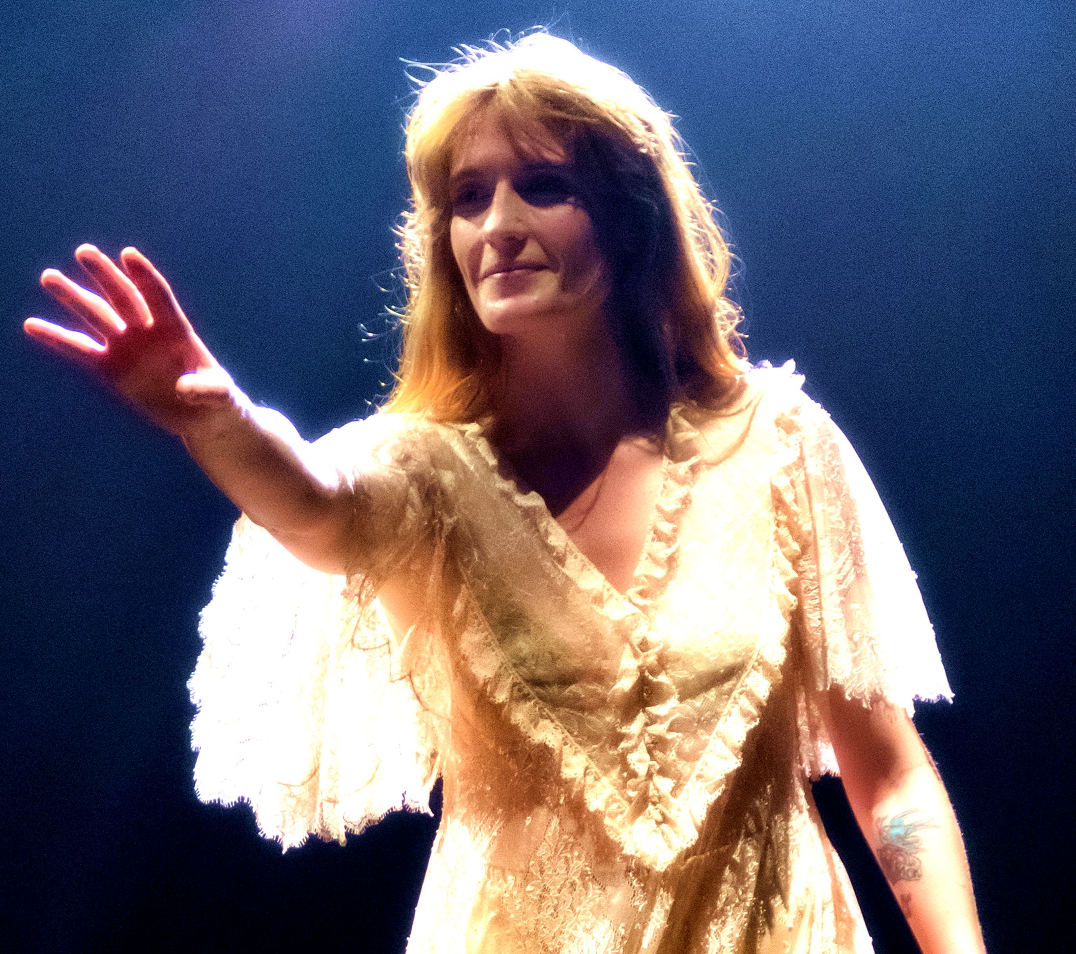 Florence Welch raises one hand towards the crowd in a cream lace dress with fluted sleeves, spotlit against a dark stage.