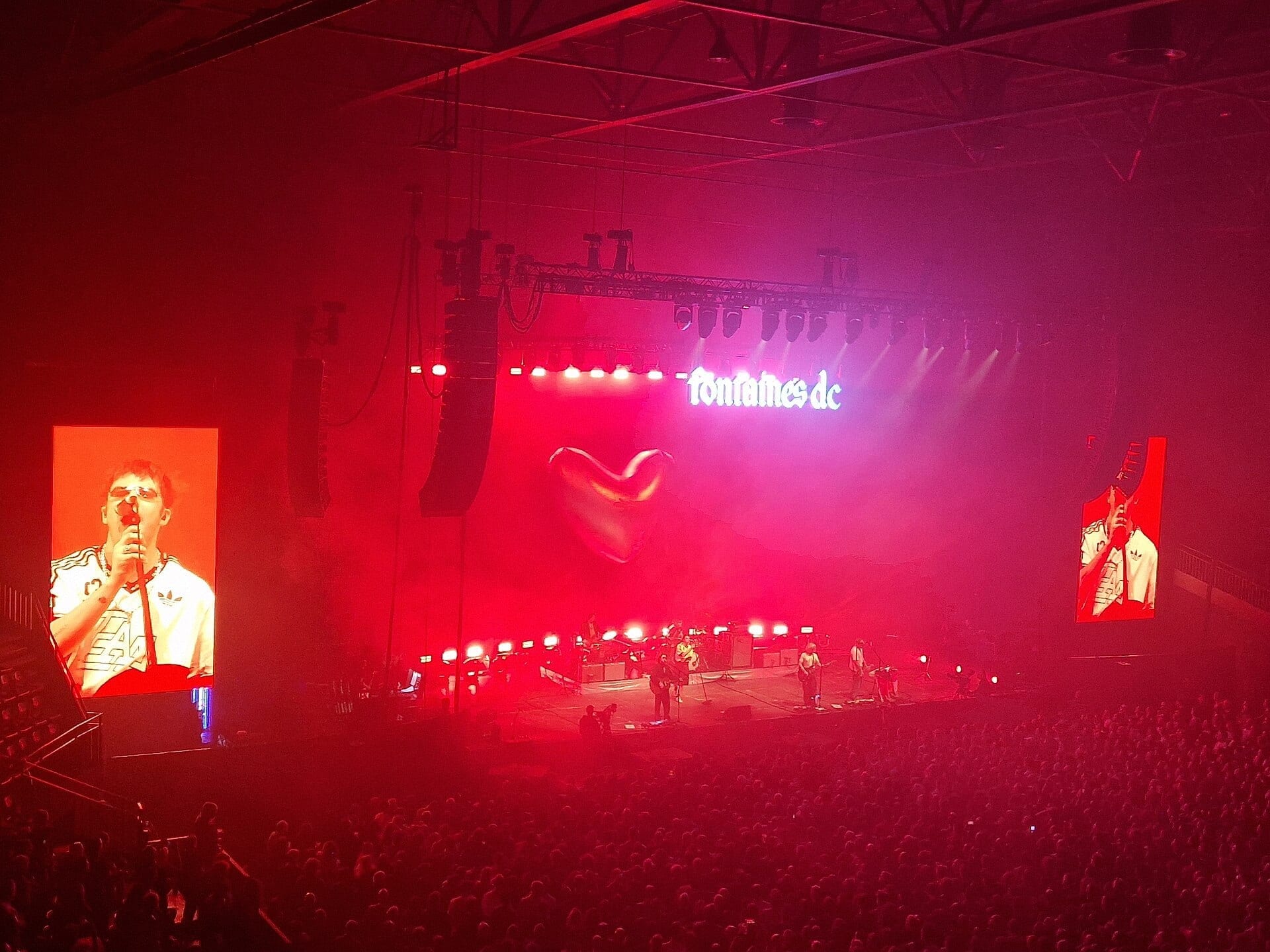 Fontaines D.C. perform in deep red light in an arena, the singer shown large on a side screen with a glowing heart above the stage.