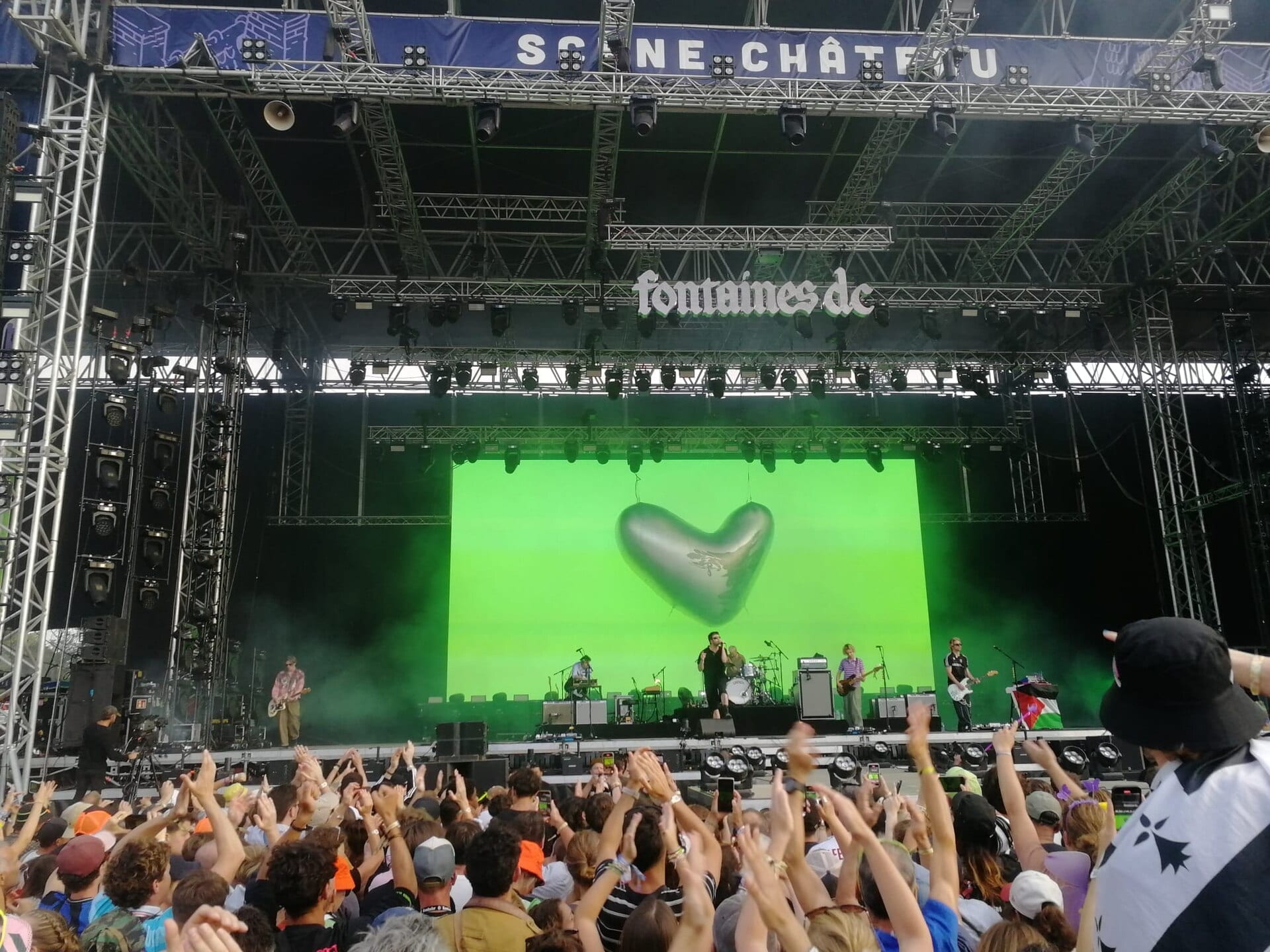 Fontaines D.C. play a festival stage washed in green light under their band name and a silver inflatable heart, with the crowd's arms up in the foreground.
