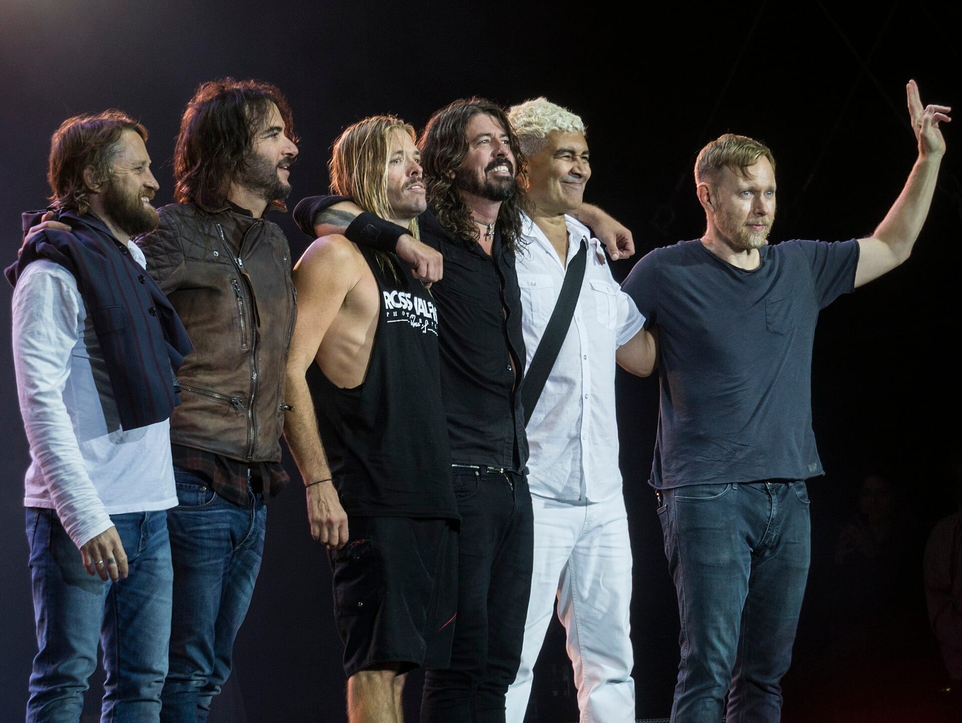The six members of Foo Fighters stand arm in arm on a dark stage, Dave Grohl at center, one member in a white shirt waving to the crowd.