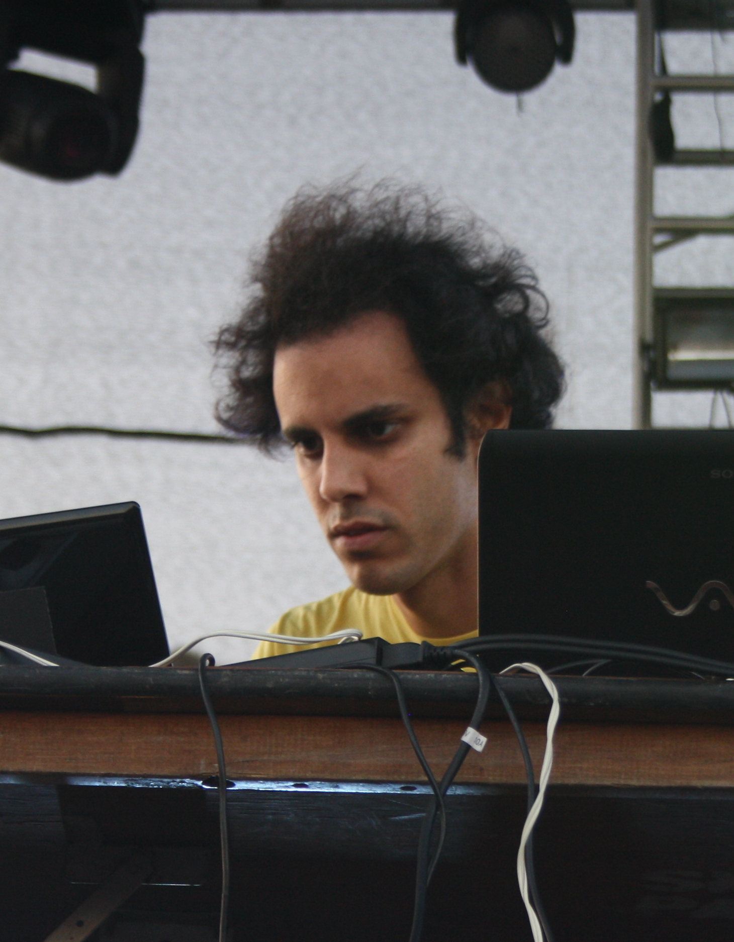 Kieran Hebden works over two laptops and a controller at a festival, curly hair falling forward, in a plain dark shirt.