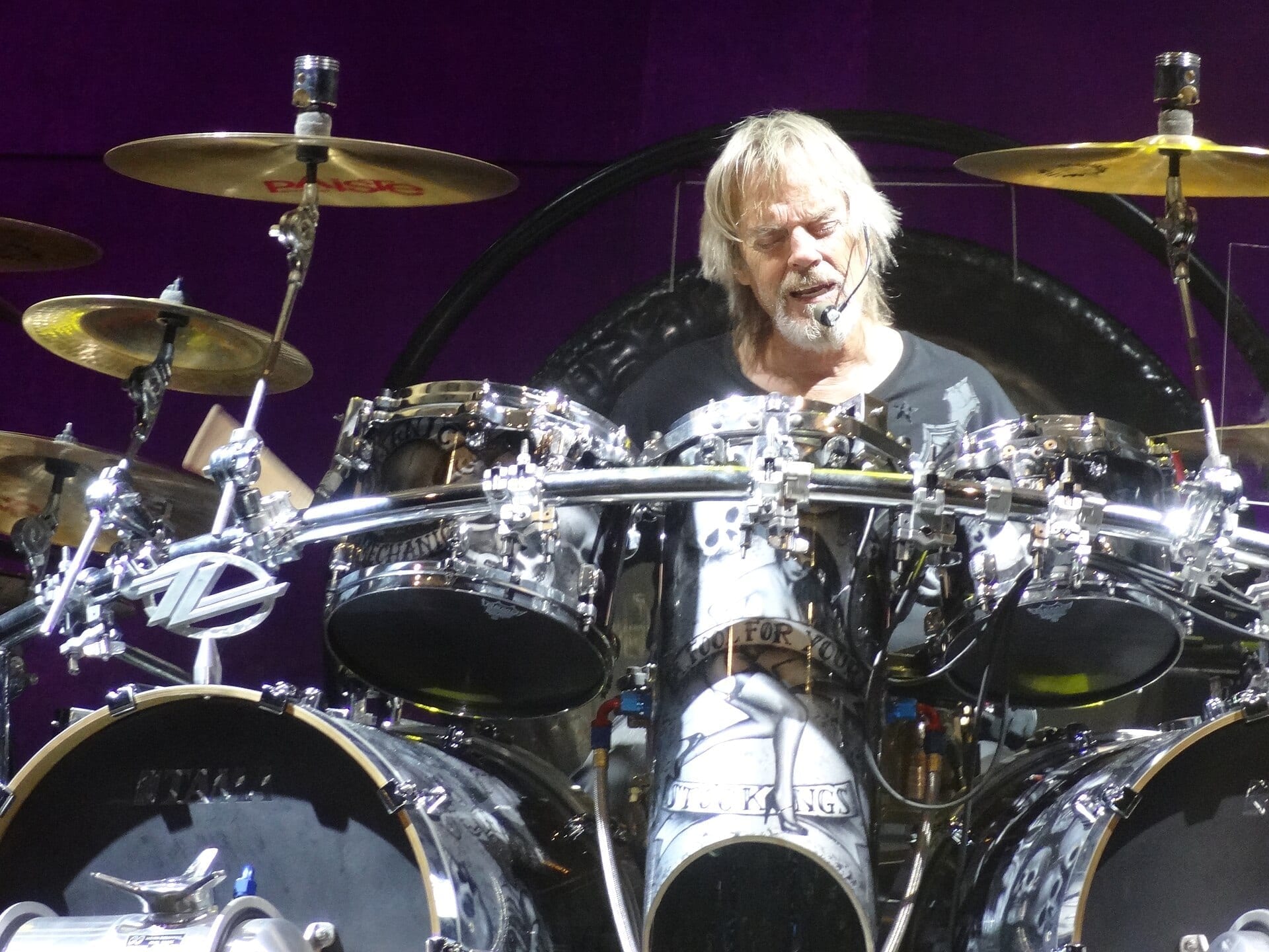 Frank Beard, with long blond hair and a white beard, plays behind his ZZ Top drum kit with the band's logo on a cymbal stand.