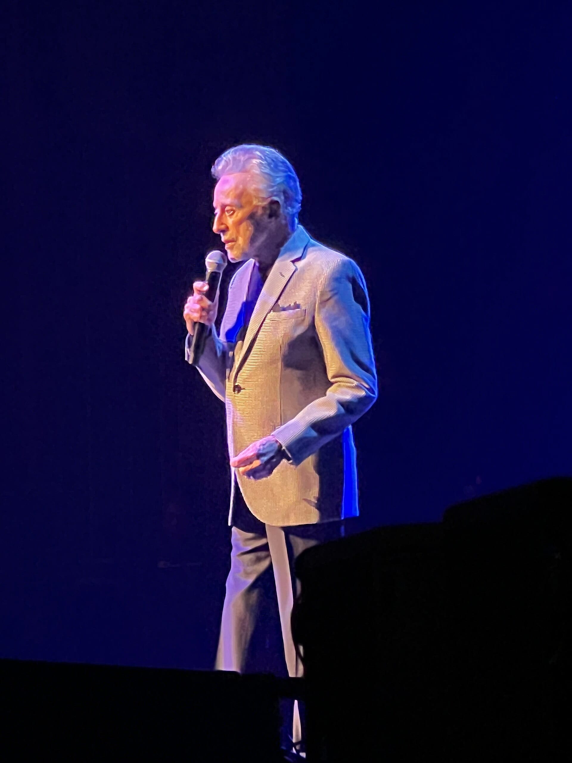 Frankie Valli sings into a handheld microphone on a dark stage lit in blue, wearing a light gold jacket.