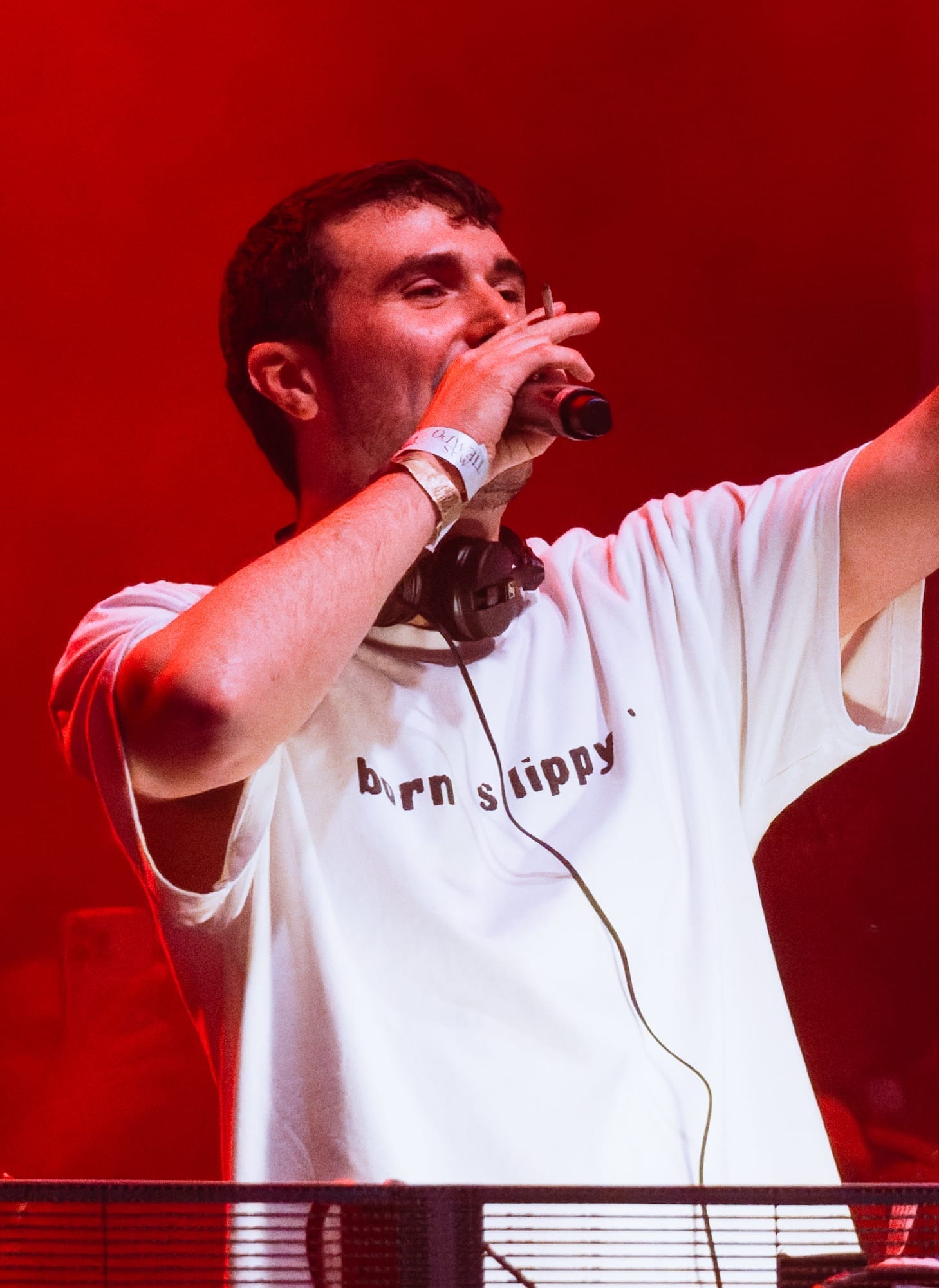 Fred again.. holding a microphone to his mouth and raising his other arm while performing in a white T-shirt reading 'born slippy'.
