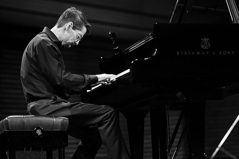 Fred Hersch, seen in profile in black and white, plays a Steinway grand piano on a dark stage.