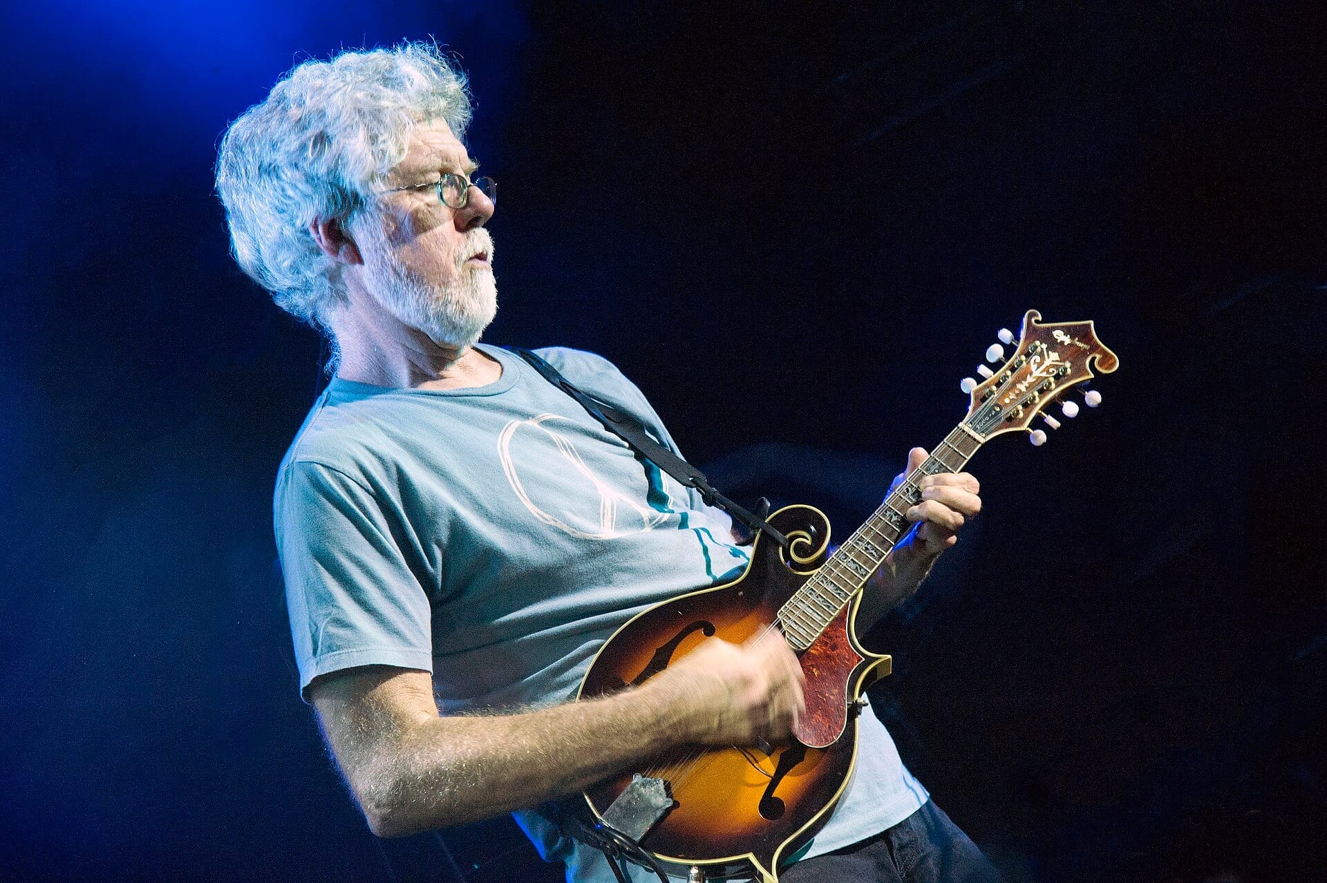 Fred Tackett plays a sunburst mandolin on a blue-lit stage, white hair and beard, wire-rimmed glasses, in a pale blue T-shirt.