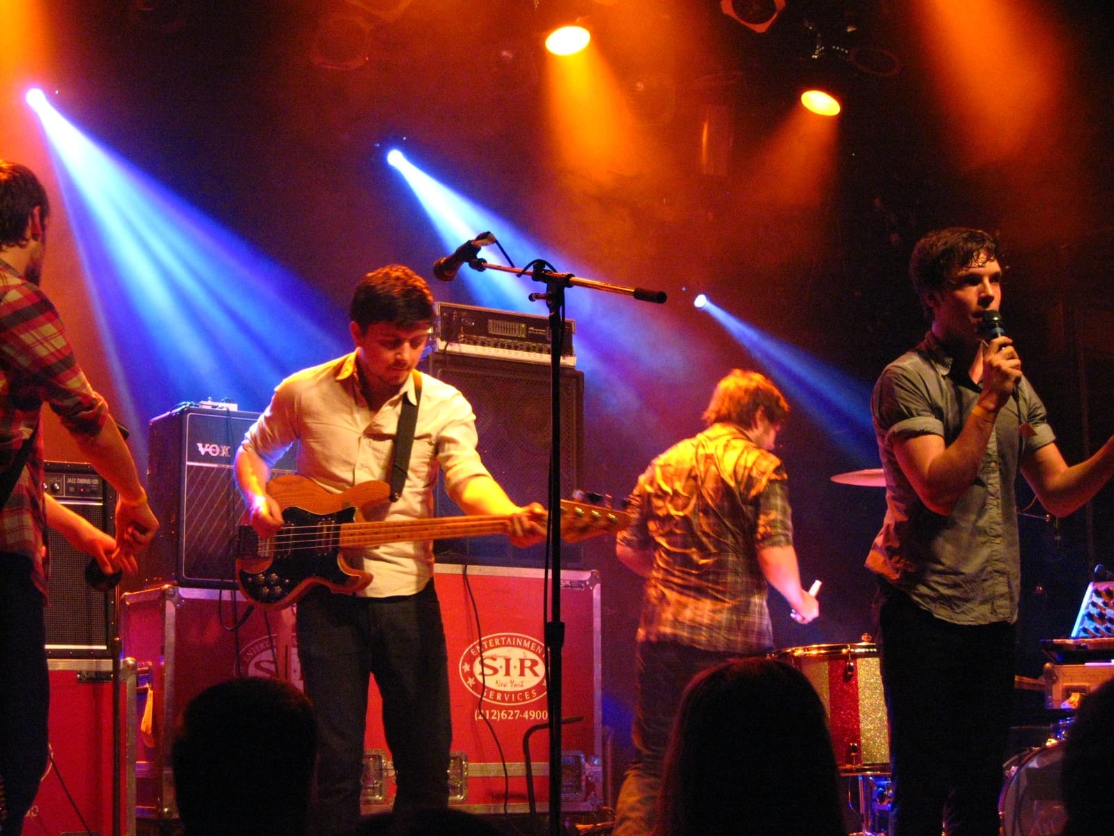 Friendly Fires play a small club stage under orange and blue lights, the singer at the right holding a microphone while a bassist works a Vox stack behind him.