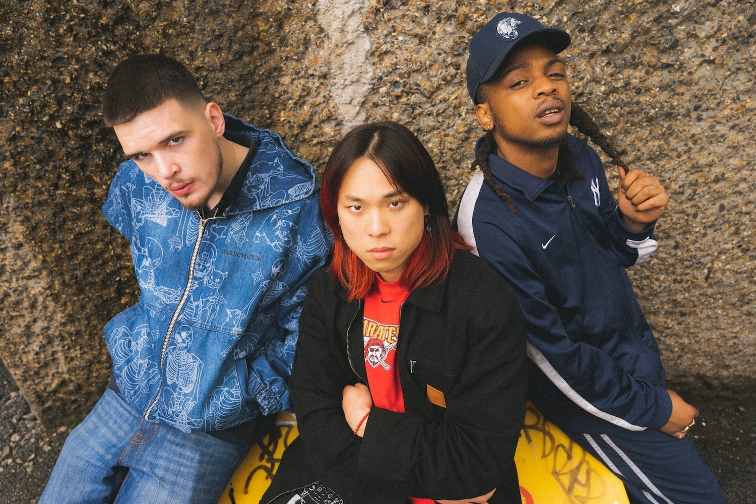 The three members of the British hip-hop group Frozemode pose together outside against a textured concrete wall.
