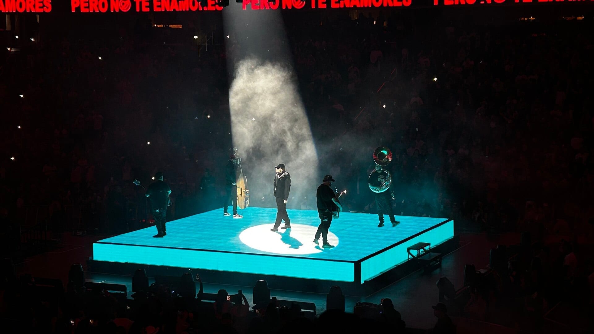 Fuerza Regida performs on a glowing teal stage under a spotlight, band members in dark clothing including a guitarist, bassist and tuba player.