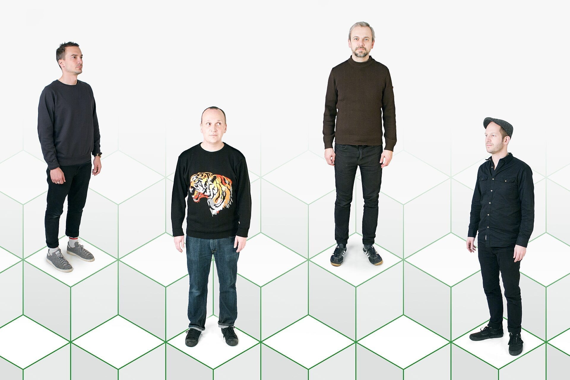 The four members of Fujiya and Miyagi stand on a stylized green hexagon-patterned floor against a white backdrop, dressed in dark casual clothes.