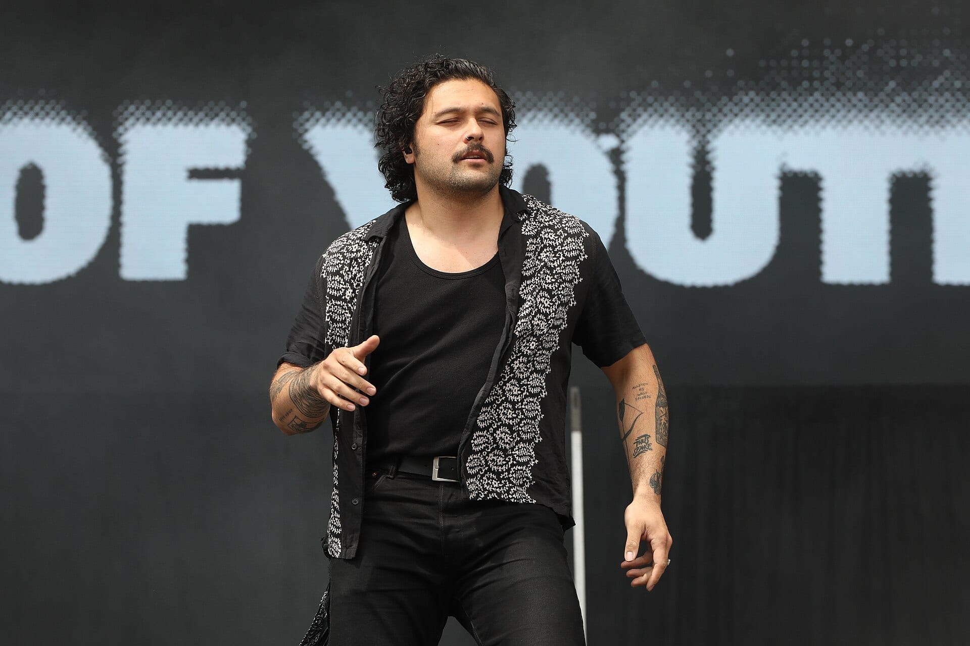 Gang of Youths frontman Dave Le'aupepe, with curly dark hair and a mustache, sings with his eyes closed in a patterned black shirt in front of a large video screen reading the band's name.
