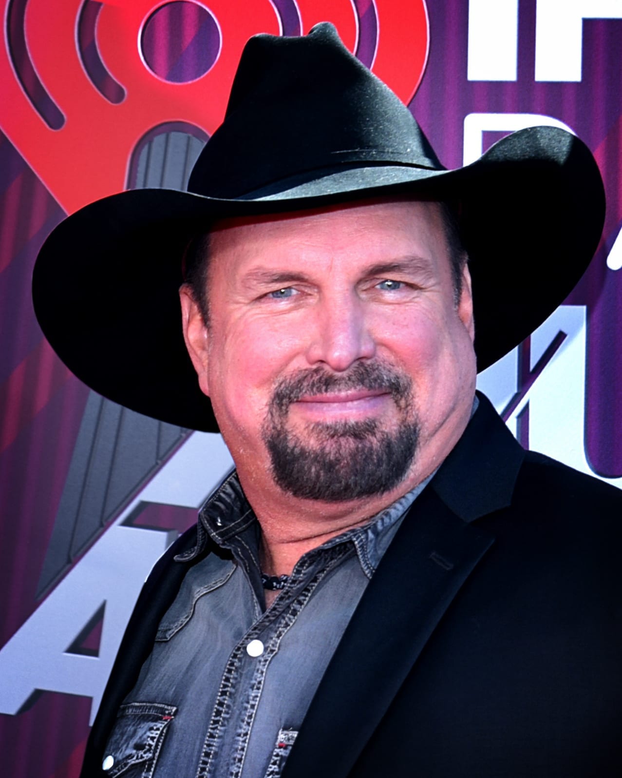 Garth Brooks smiles for the camera at a red carpet event, wearing a black wide-brim cowboy hat, a gray denim snap shirt, and a black blazer, with a red iHeartRadio backdrop behind him.
