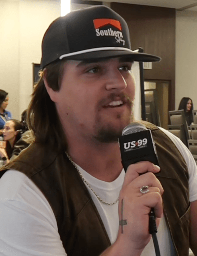 Gavin Adcock, wearing a backwards trucker cap and a gold chain, speaks into a microphone branded US 99 during an interview.