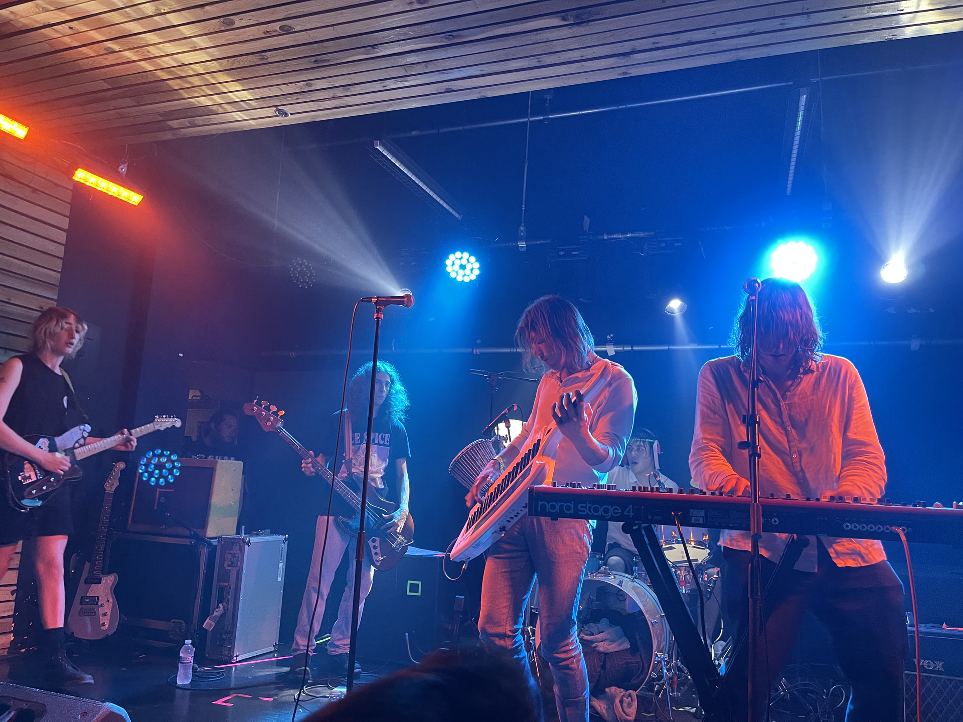 Five members of Geese play a small club stage under blue and orange light, one on a white keytar and another leaning over a stage piano.