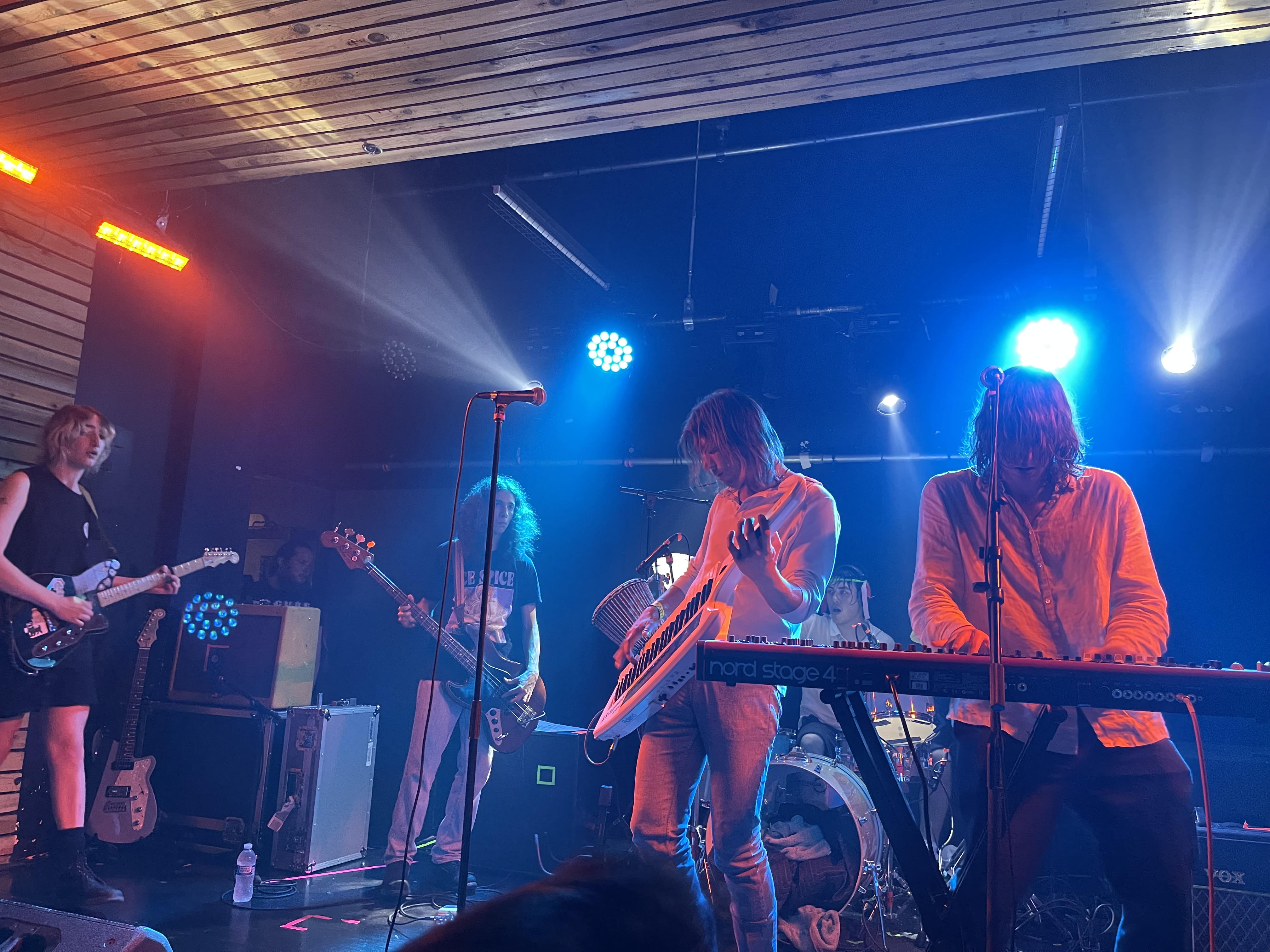 Five members of Geese play a small club stage lit blue and orange, a bassist and guitarists to the left and the singer at a keyboard on the right.