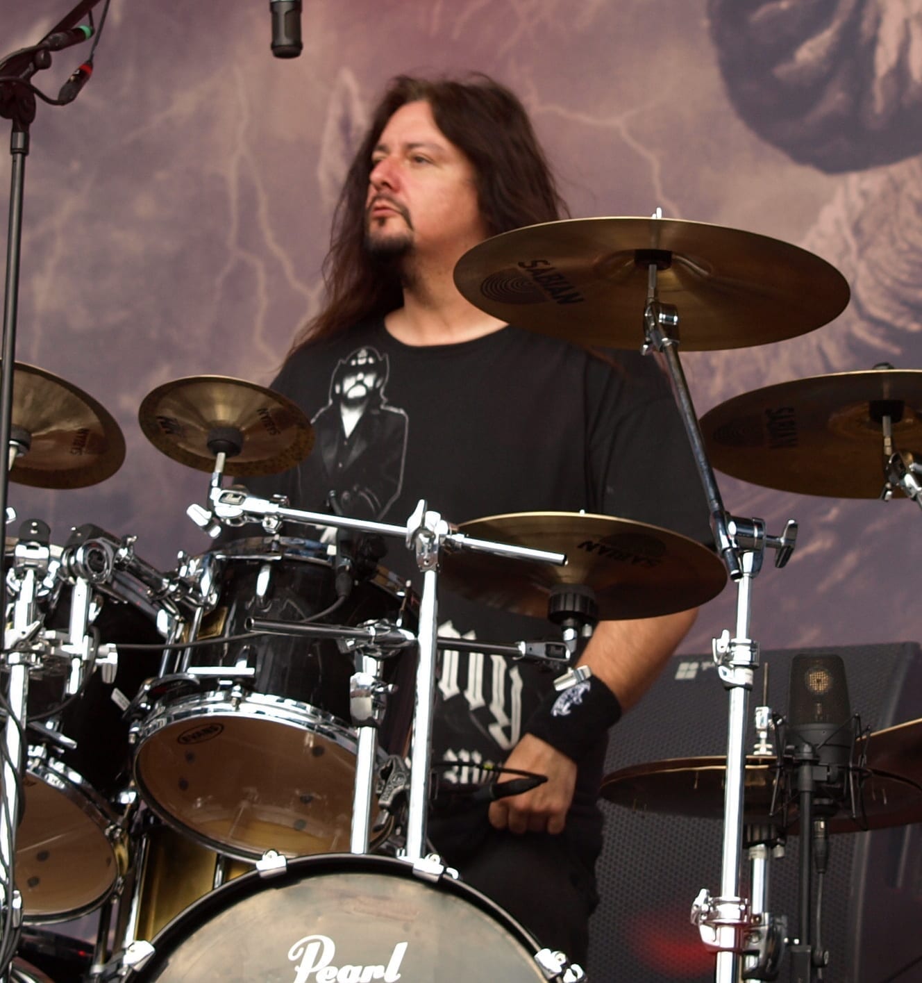 Gene Hoglan plays drums on an outdoor festival stage, wearing a black t-shirt with a portrait graphic, his long hair mid-motion.