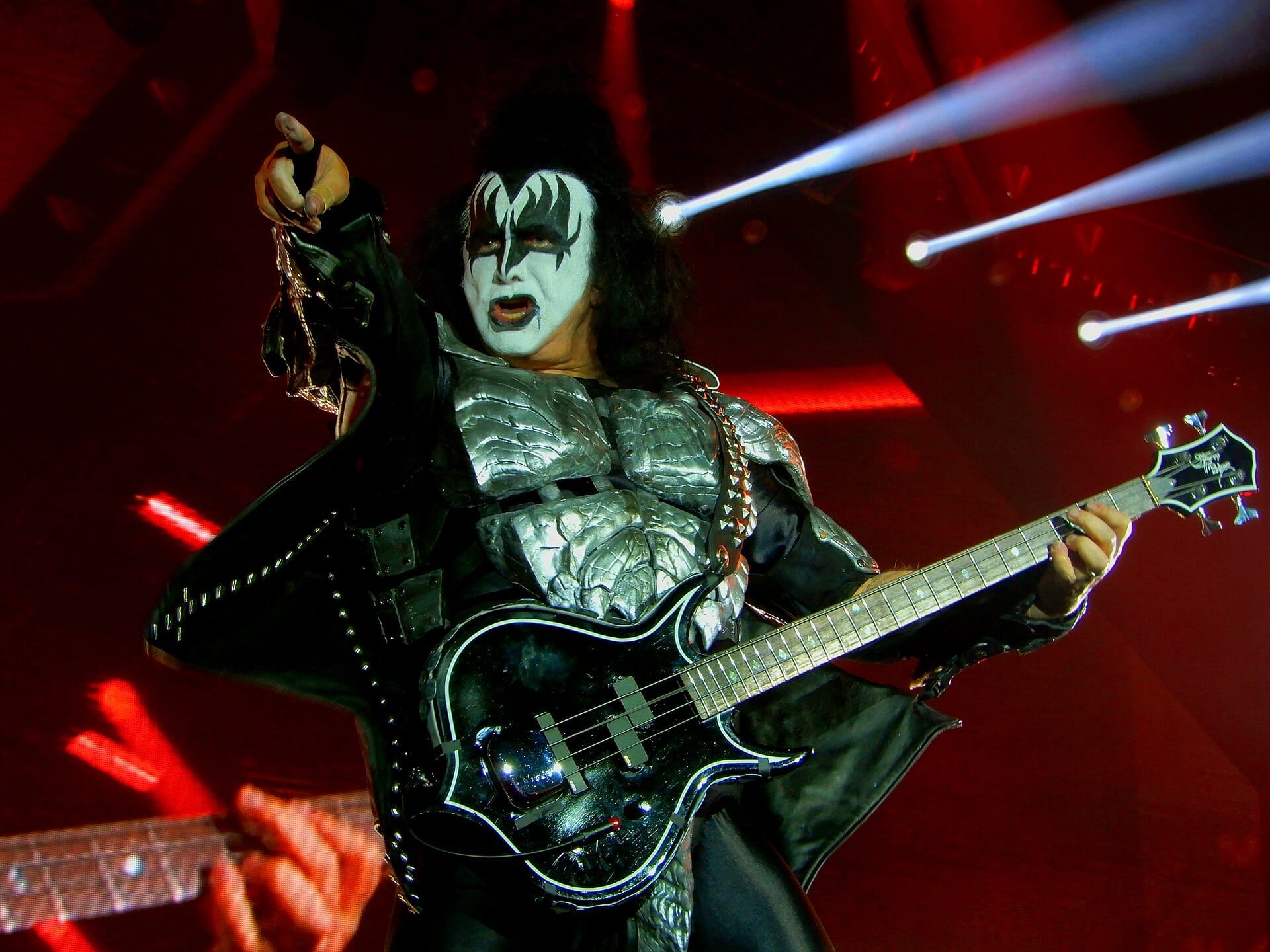 Gene Simmons in full Demon makeup and silver armour points into the crowd from a red-lit stage, holding his axe-shaped bass.