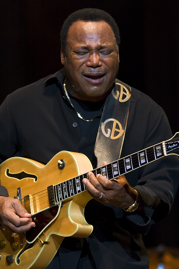 A guitarist in a black shirt plays a hollow-body archtop guitar with his eyes shut and mouth open, a strap lettered GB across his chest