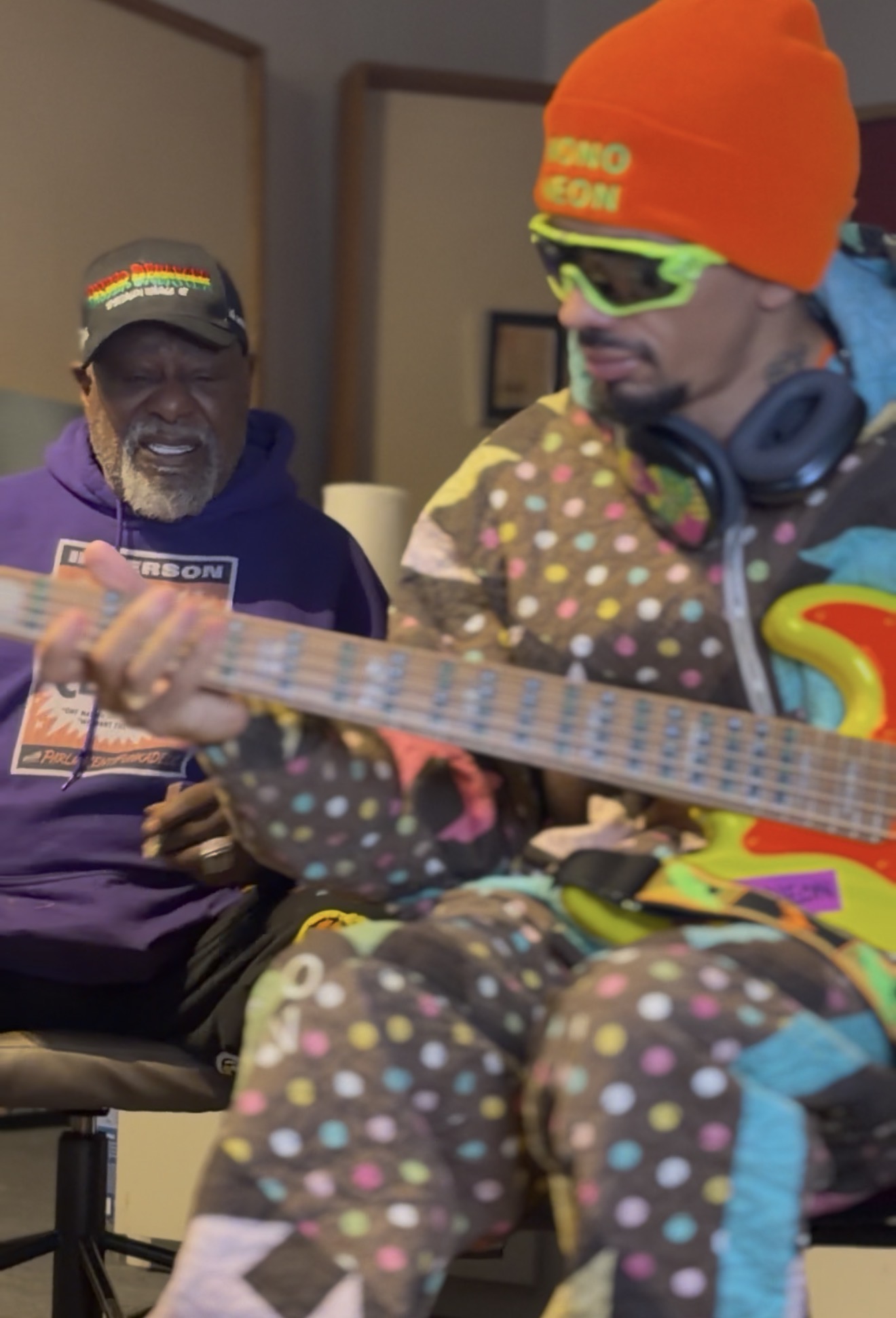 George Clinton, white-bearded and in a purple hooded top, stands beside the bassist MonoNeon, who wears an orange beanie printed with his name.