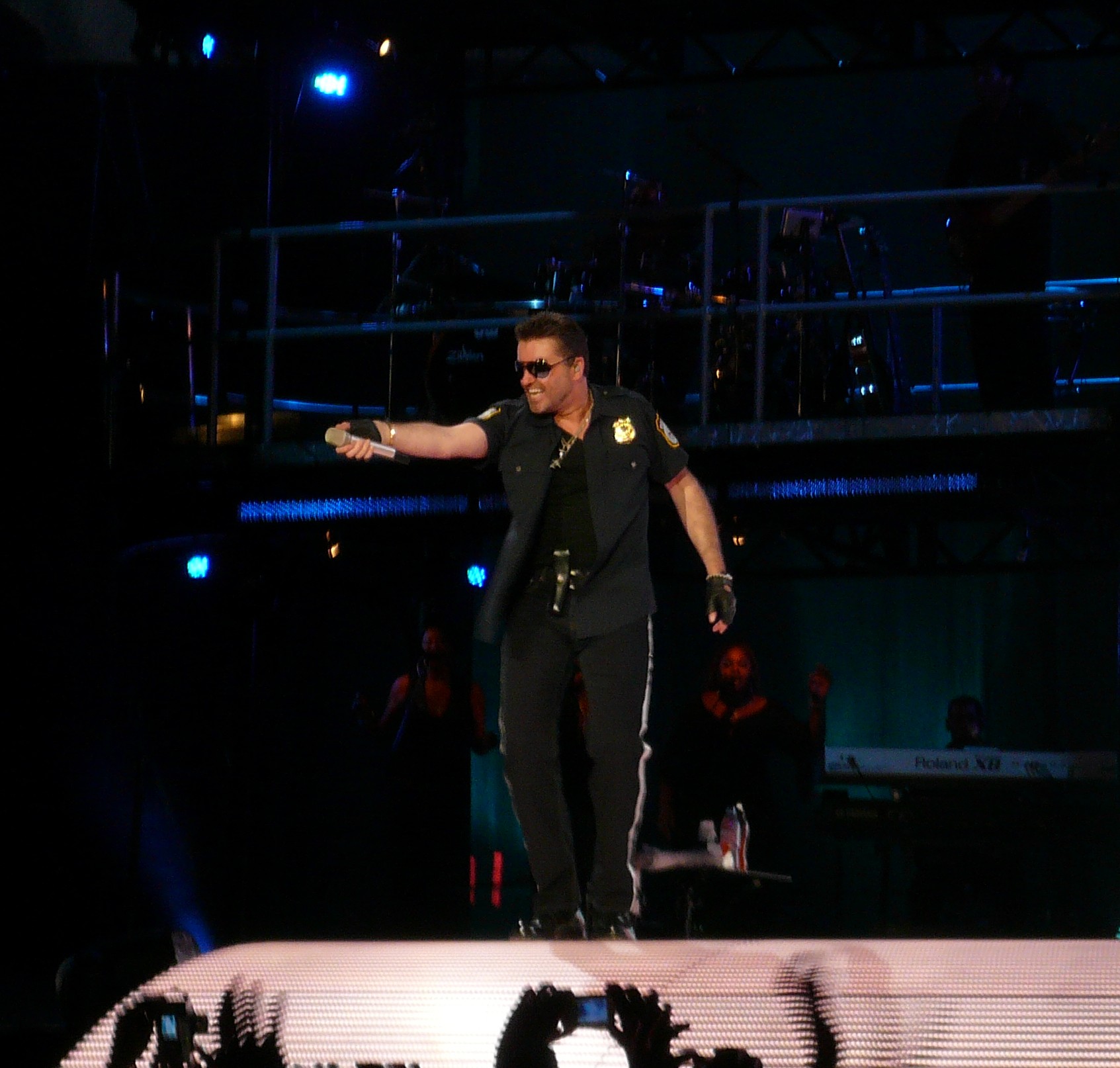 George Michael strides across a stadium stage in sunglasses and a police-style shirt, arm extended with a microphone.