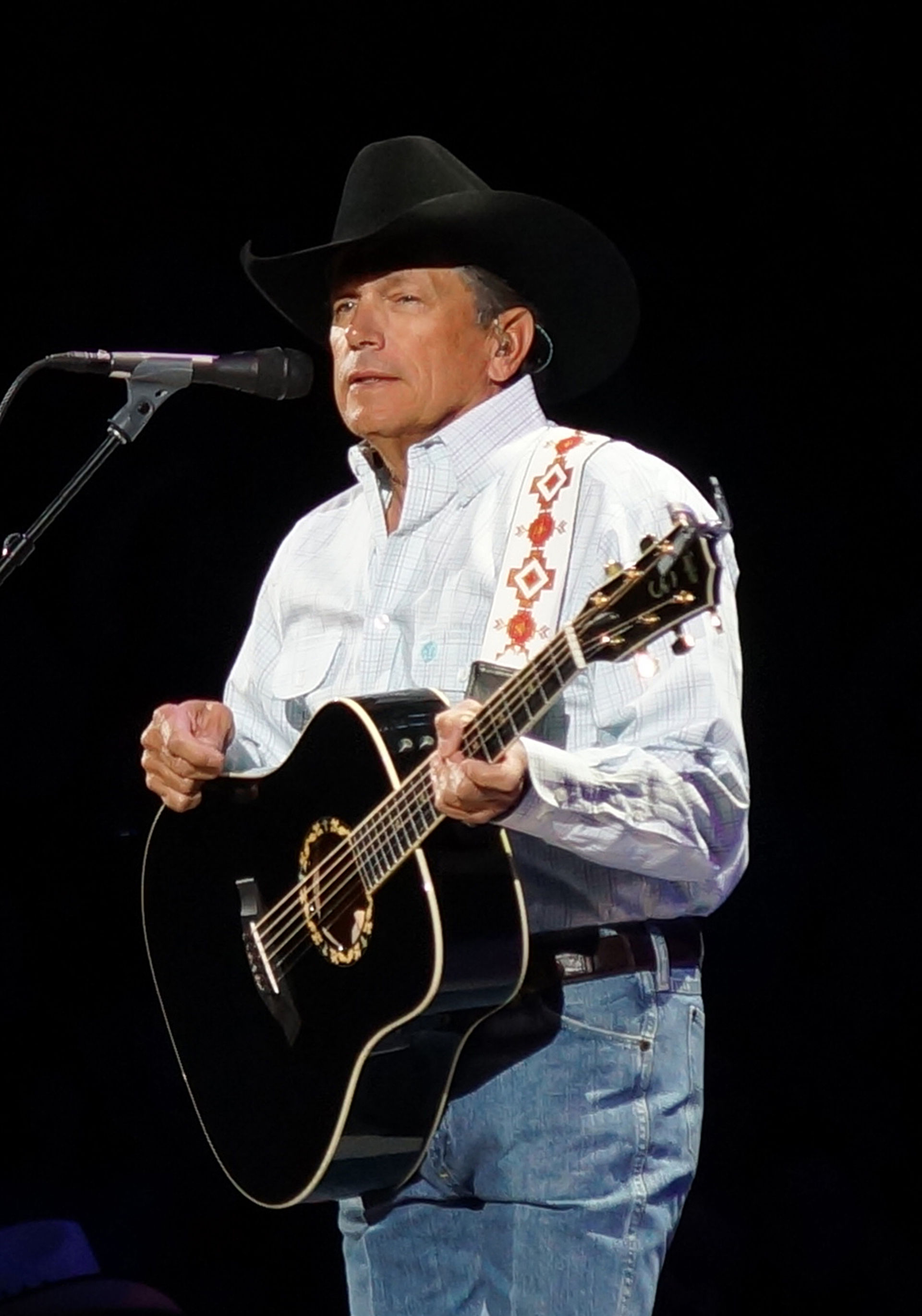 George Strait stands at a microphone with a black acoustic guitar, in a black cowboy hat, pale plaid shirt and jeans against a dark stage.
