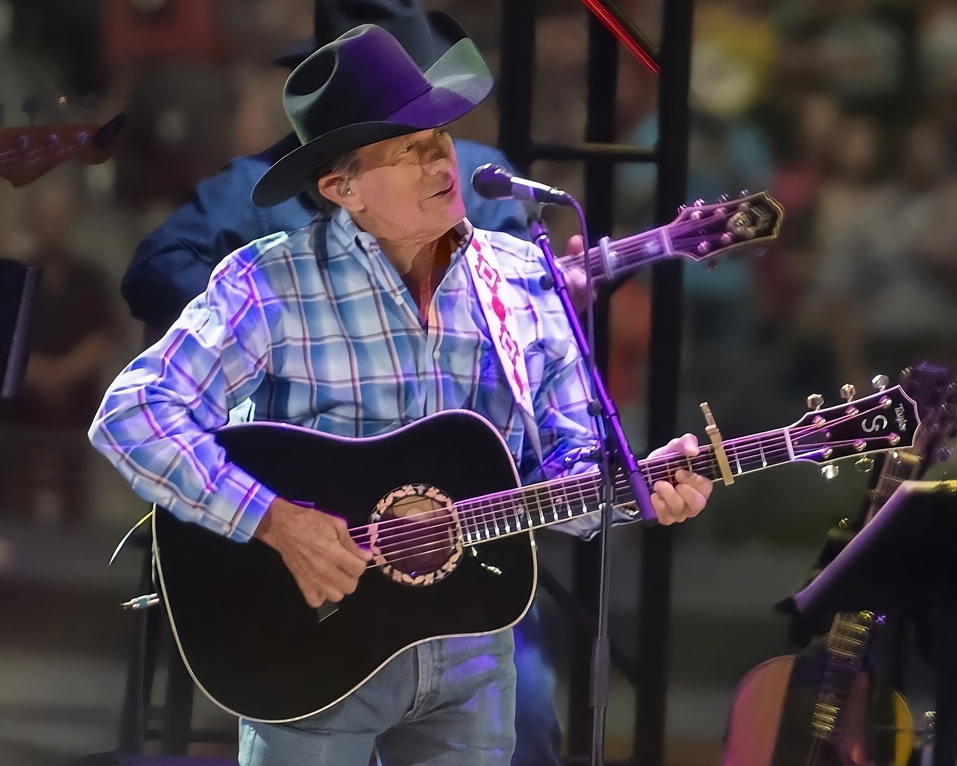 George Strait plays an acoustic guitar and leans into a microphone in a black cowboy hat and blue plaid shirt, a packed stadium behind him.