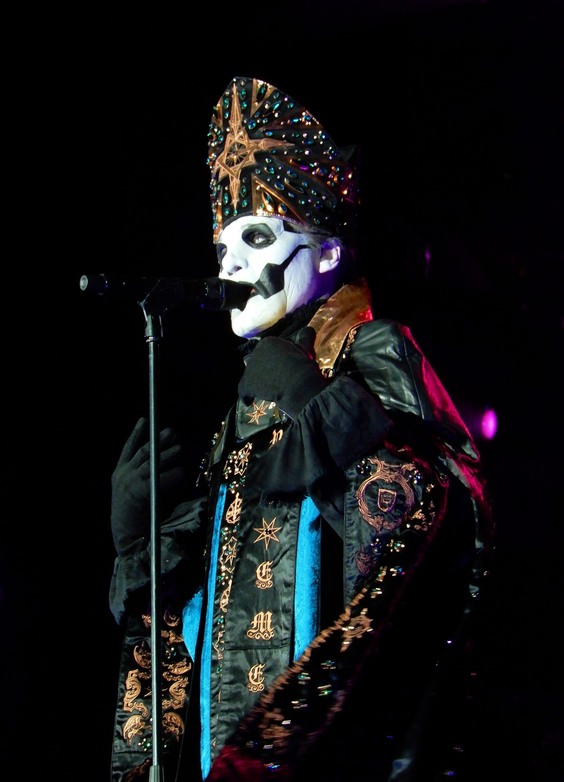 Tobias Forge in full Ghost stage makeup and a dark ornate costume, singing into a handheld microphone.