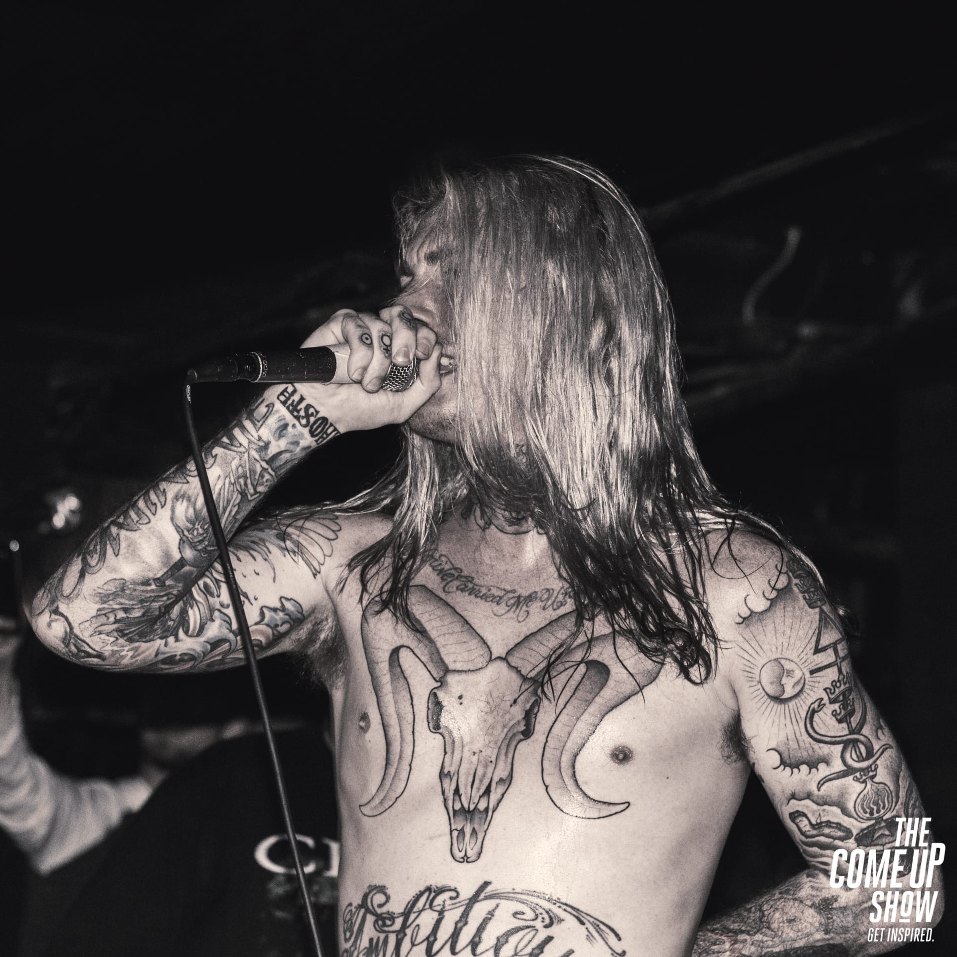 Ghostemane screams into a microphone on stage, shirtless and heavily tattooed with long hair covering his face, in a black and white photo.