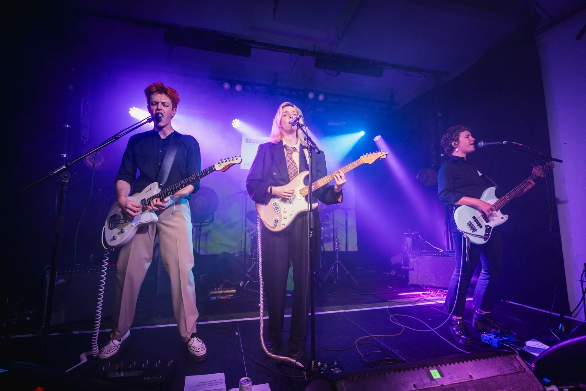 Gia Ford singing and playing guitar on stage with her band, flanked by a guitarist and bassist, under purple stage lighting at a club show.