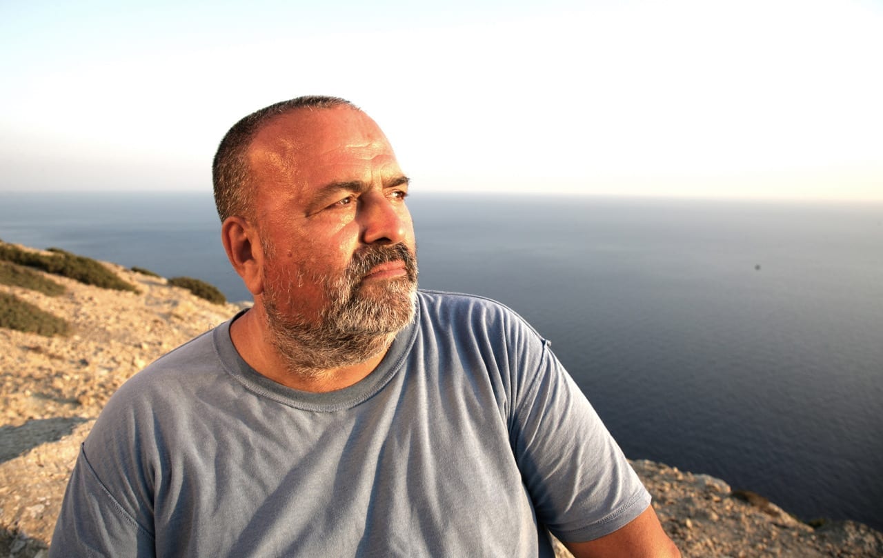 A close-up portrait of Gigi Masin looking out over the ocean from a rocky cliff, wearing a gray T-shirt.
