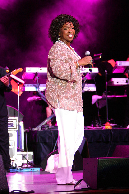 Gladys Knight sings into a handheld microphone in a pink patterned jacket and white trousers, her band and keyboards behind her.