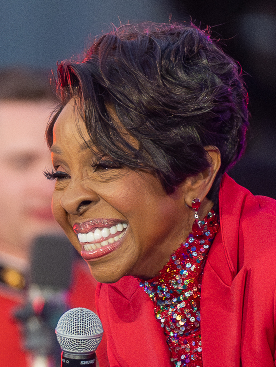 Gladys Knight smiles mid-song in a bright red blazer, holding a microphone, under warm stage light.