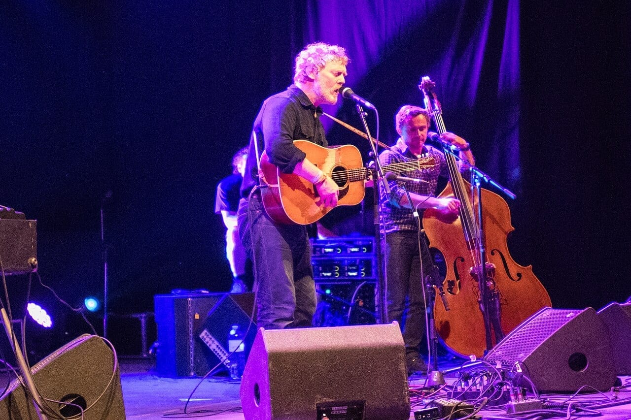 Glen Hansard sings into a microphone under purple stage light while playing an acoustic guitar, with a double bass player behind him.