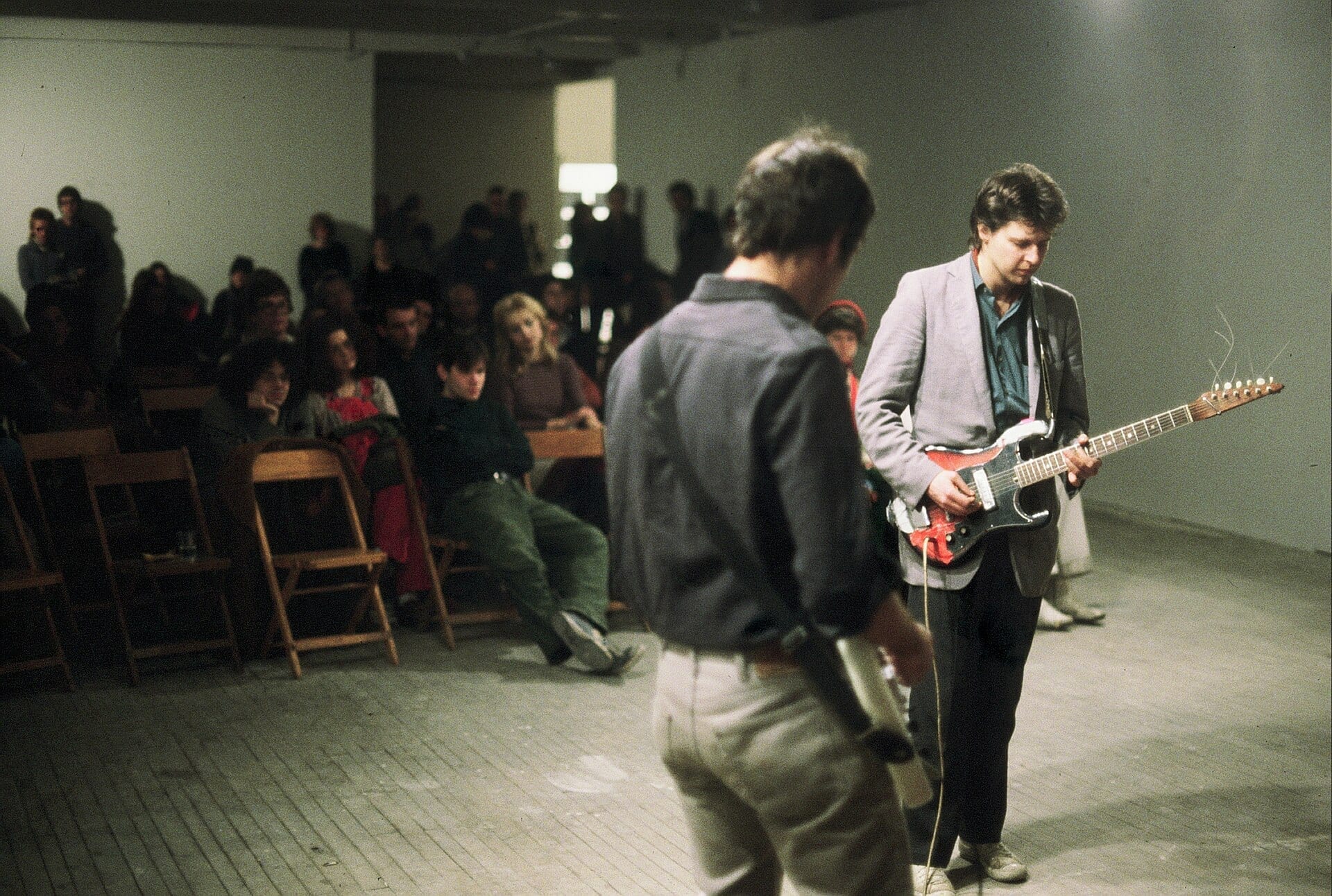 Glenn Branca plays an electric guitar in a bare loft space before a small seated audience, another musician standing nearby with a handheld device.