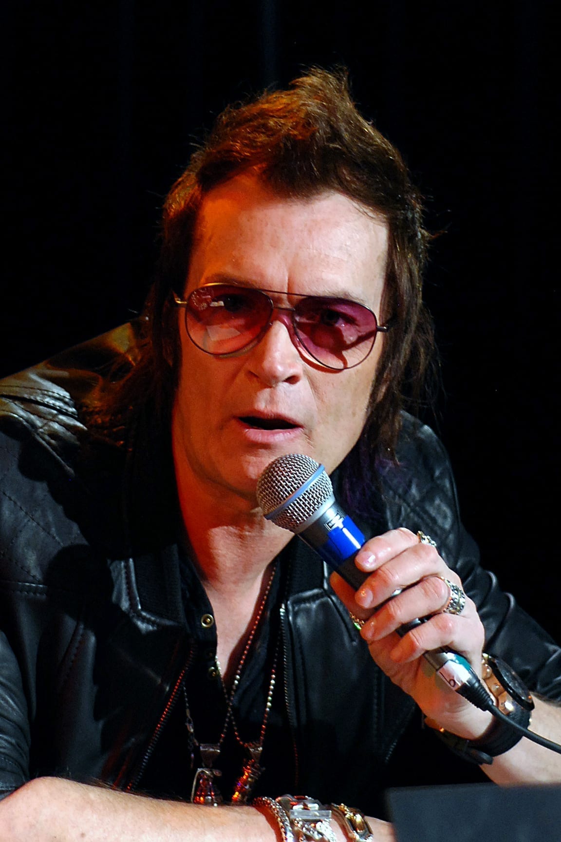 Glenn Hughes, wearing tinted aviator sunglasses and a black leather jacket, speaks into a microphone he holds close to his mouth.