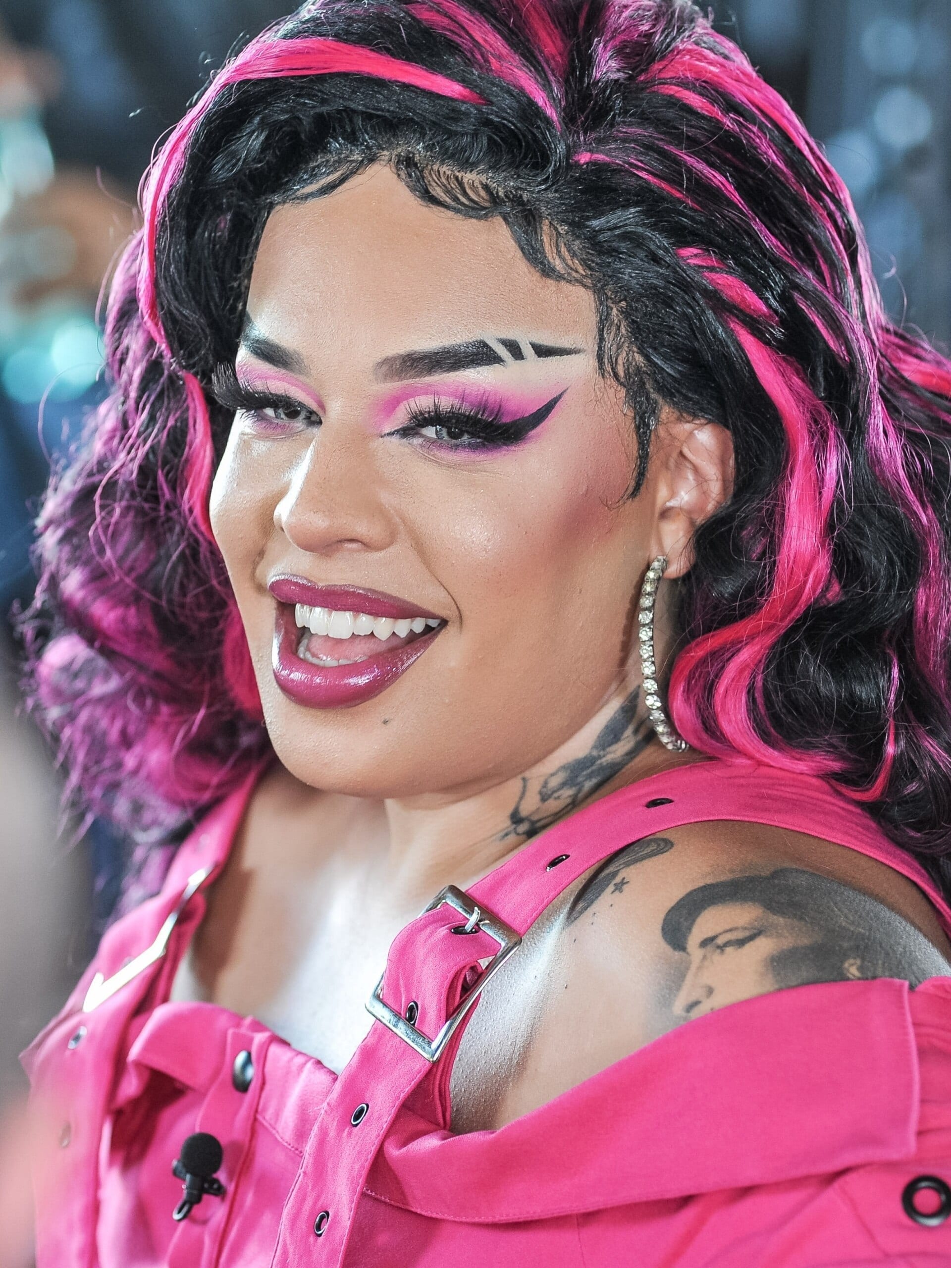 Gloria Groove smiles in close-up with bold pink and black eye makeup, pink-streaked hair, and a pink studded outfit.