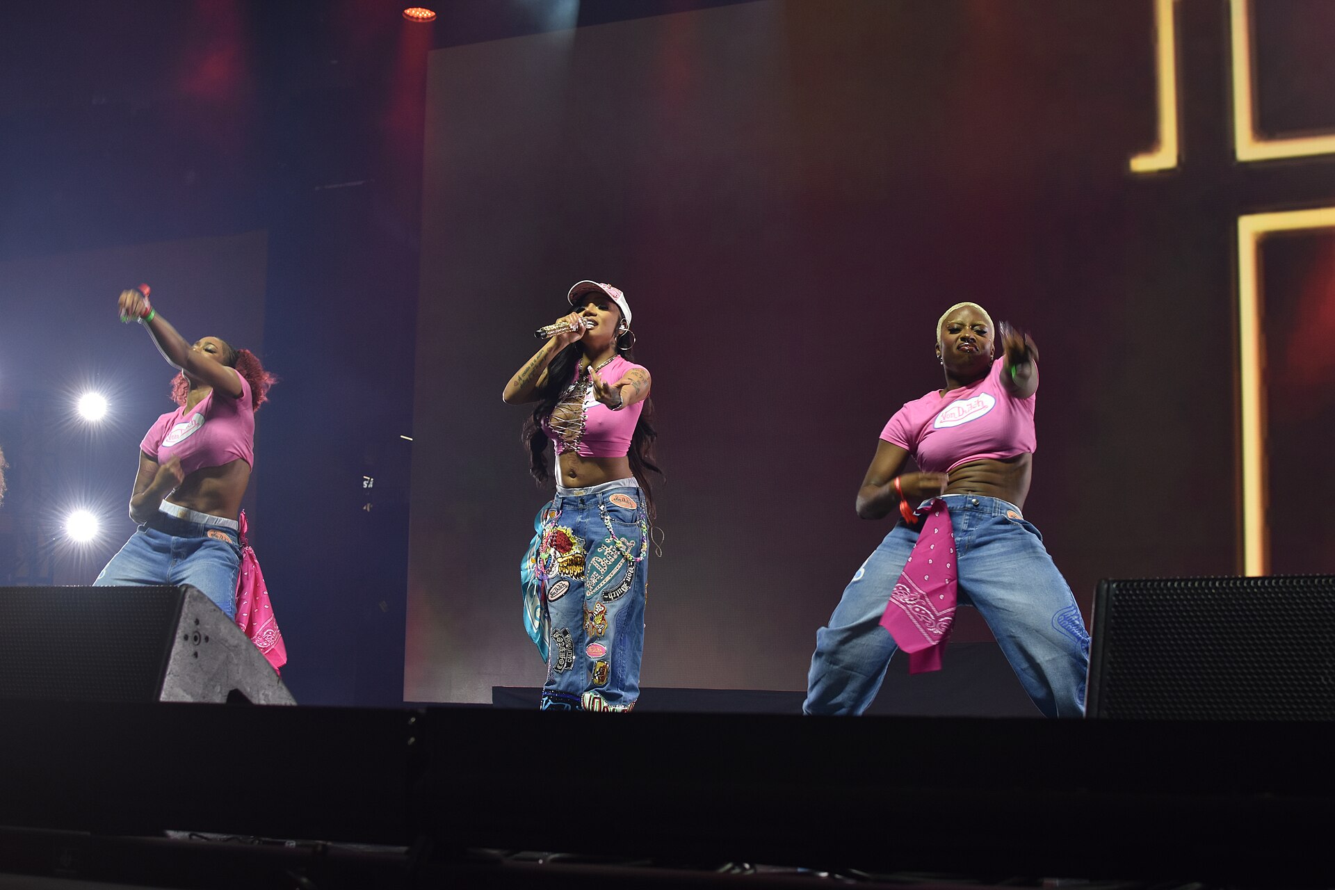 GloRilla raps into a gold microphone centre stage in a pink crop top, white cap and patched baggy jeans, flanked by two dancers dressed to match.
