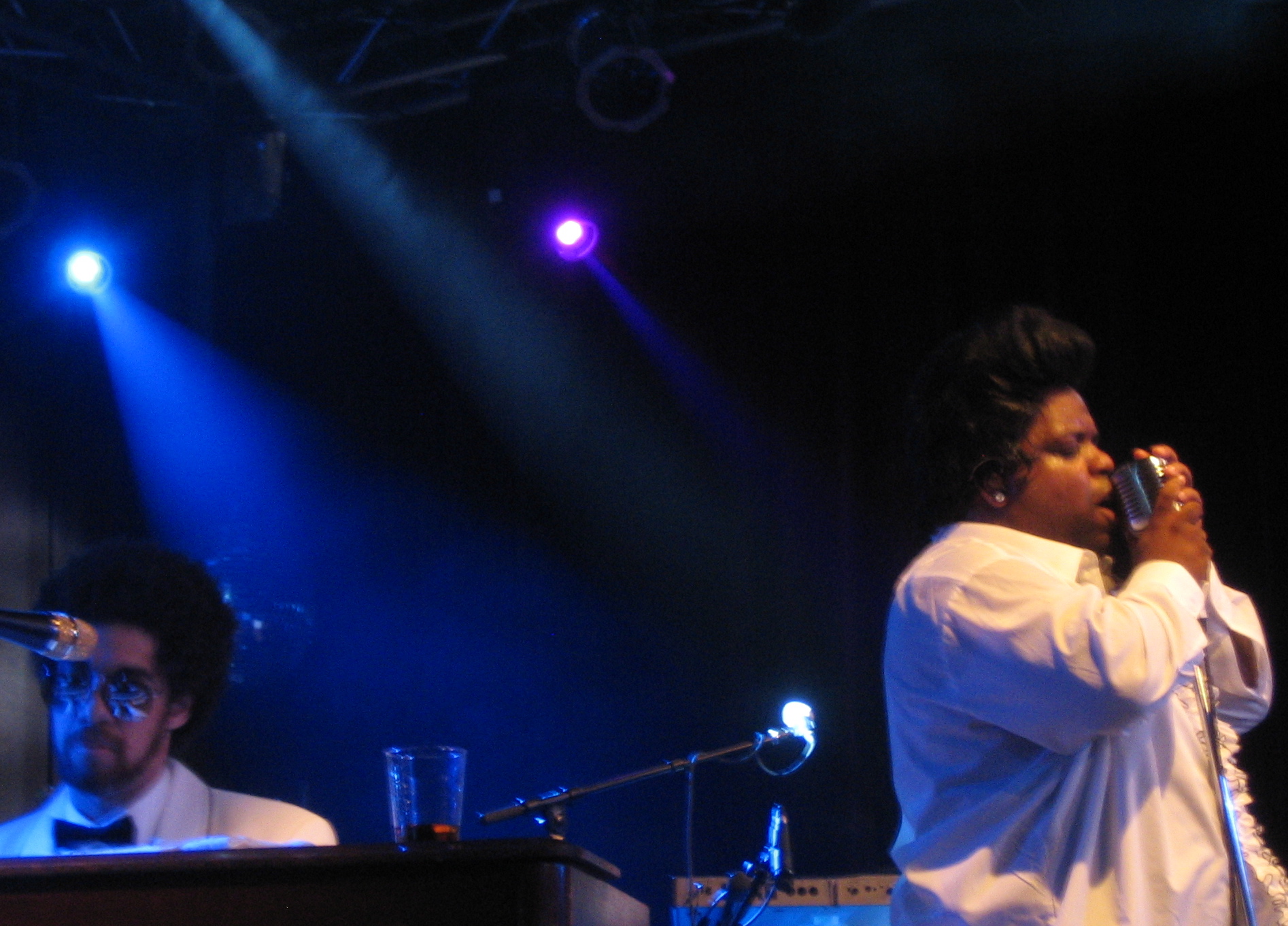 CeeLo Green singing into a vintage ribbon microphone in a white shirt on a blue-lit stage while Danger Mouse plays keyboards in a white dinner jacket and bow tie at the left