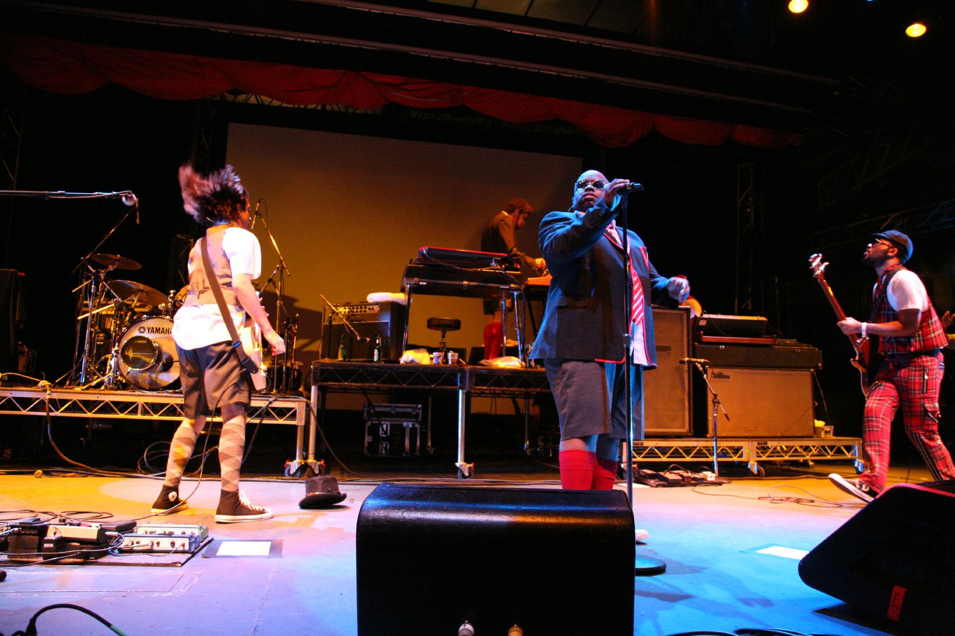 A singer in a blue uniform jacket, shorts and red socks stands at a mic stand mid-stage with a costumed backing band, guitarist and bassist on either side
