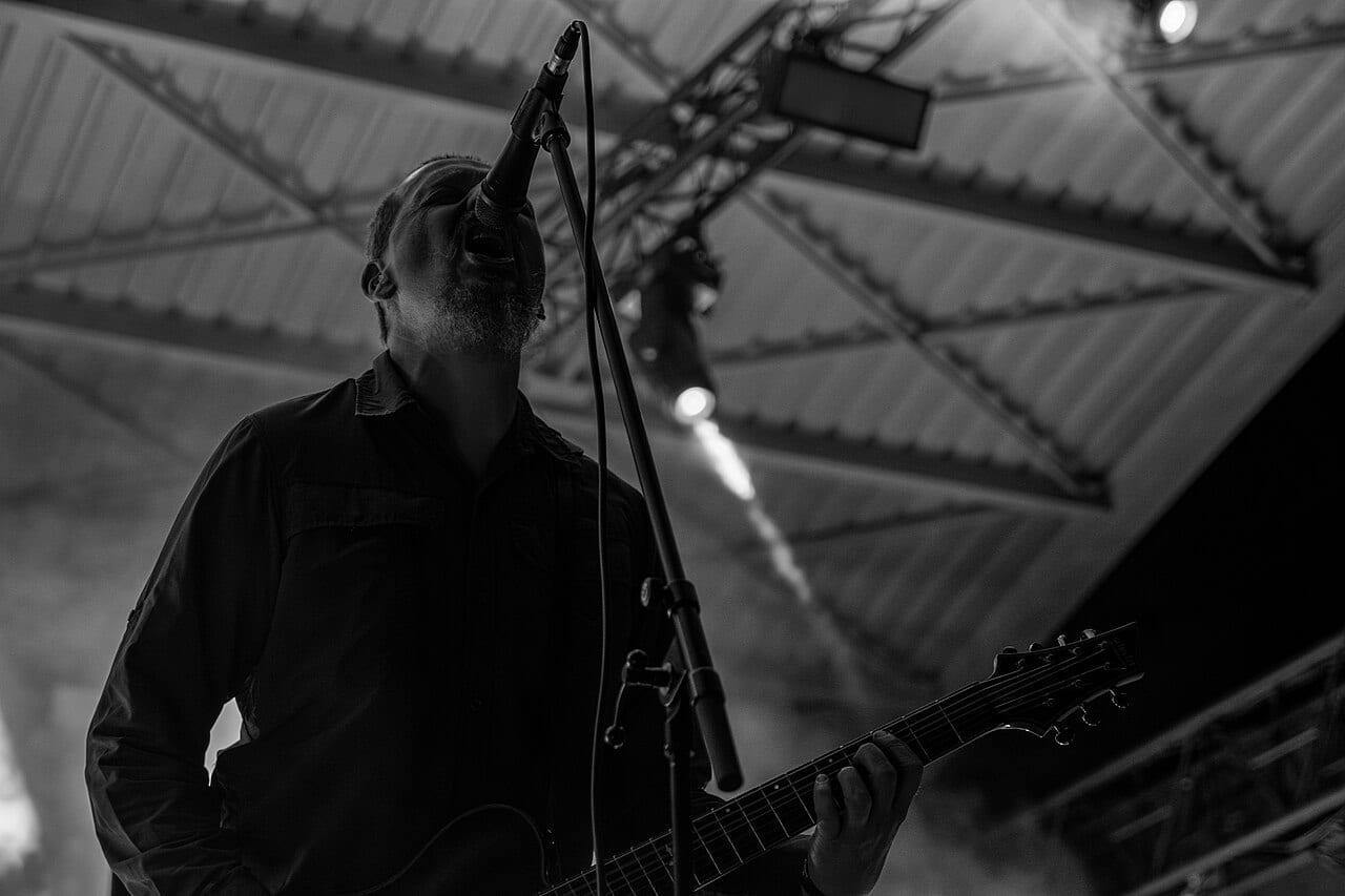 Justin Broadrick of Godflesh tilts his head back to sing into a microphone while playing guitar, in a black and white photograph.