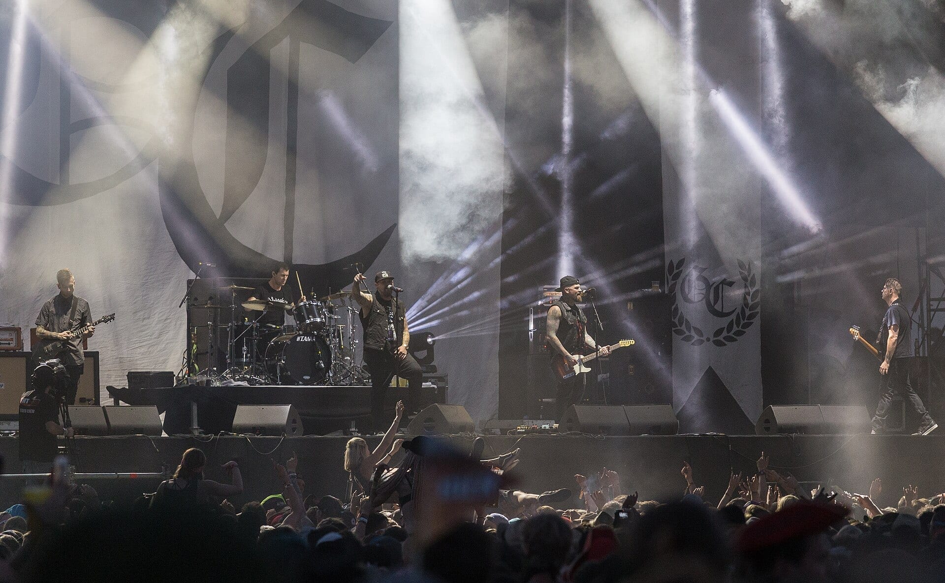 Good Charlotte perform on a smoke-lit festival stage with large GC banners behind them, as a packed crowd raises their hands in the foreground.