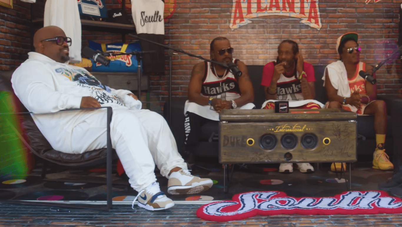 The four members of Goodie Mob sit for a backstage interview in matching Atlanta Hawks jerseys, with CeeLo Green in white at the front and a large wooden speaker cabinet between them.