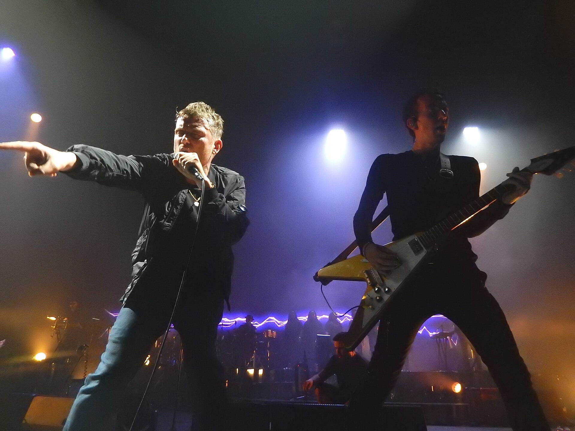 Two Gorillaz musicians performing on a dark stage under blue and purple lights, one singing into a microphone and the other playing an electric guitar.